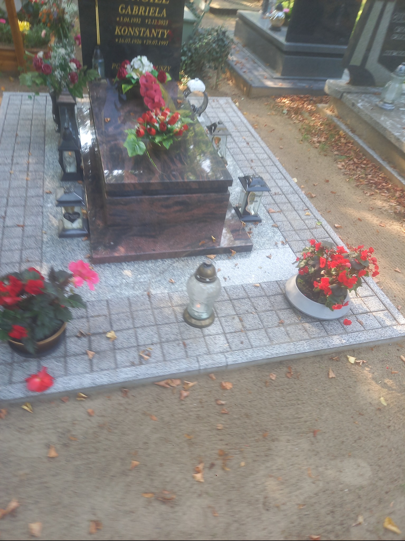 Granitowy nagrobek z kwiatami i lampionami na cmentarzu, otoczony kostką brukową i opadłymi liśćmi. Ujęcie z góry, w słoneczny dzień.