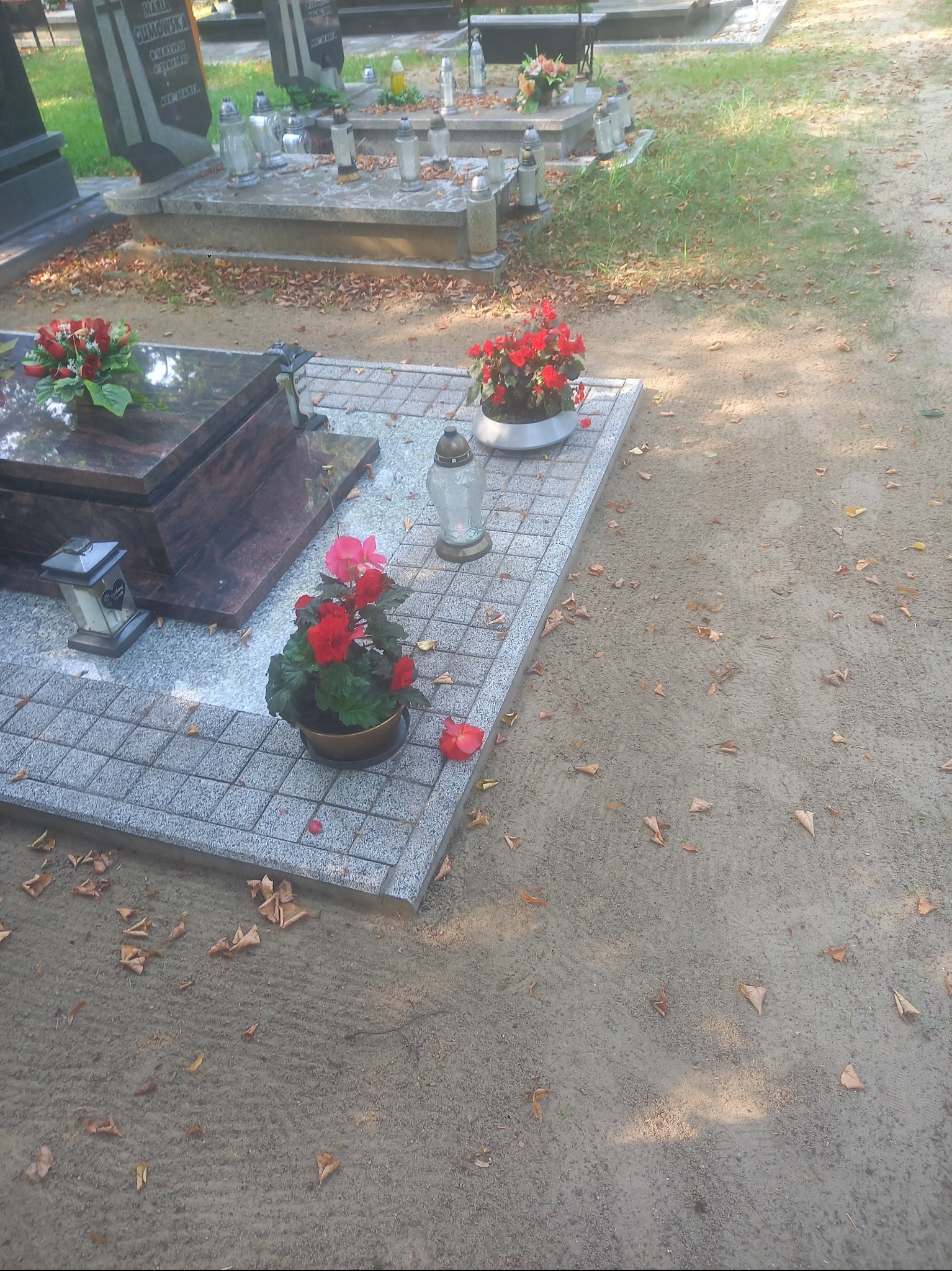 Nagrobek z kwiatami i zniczem na cmentarzu, jesienne liście na ziemi. Widok z góry, spokojna atmosfera zadumy i pamięci o zmarłych.