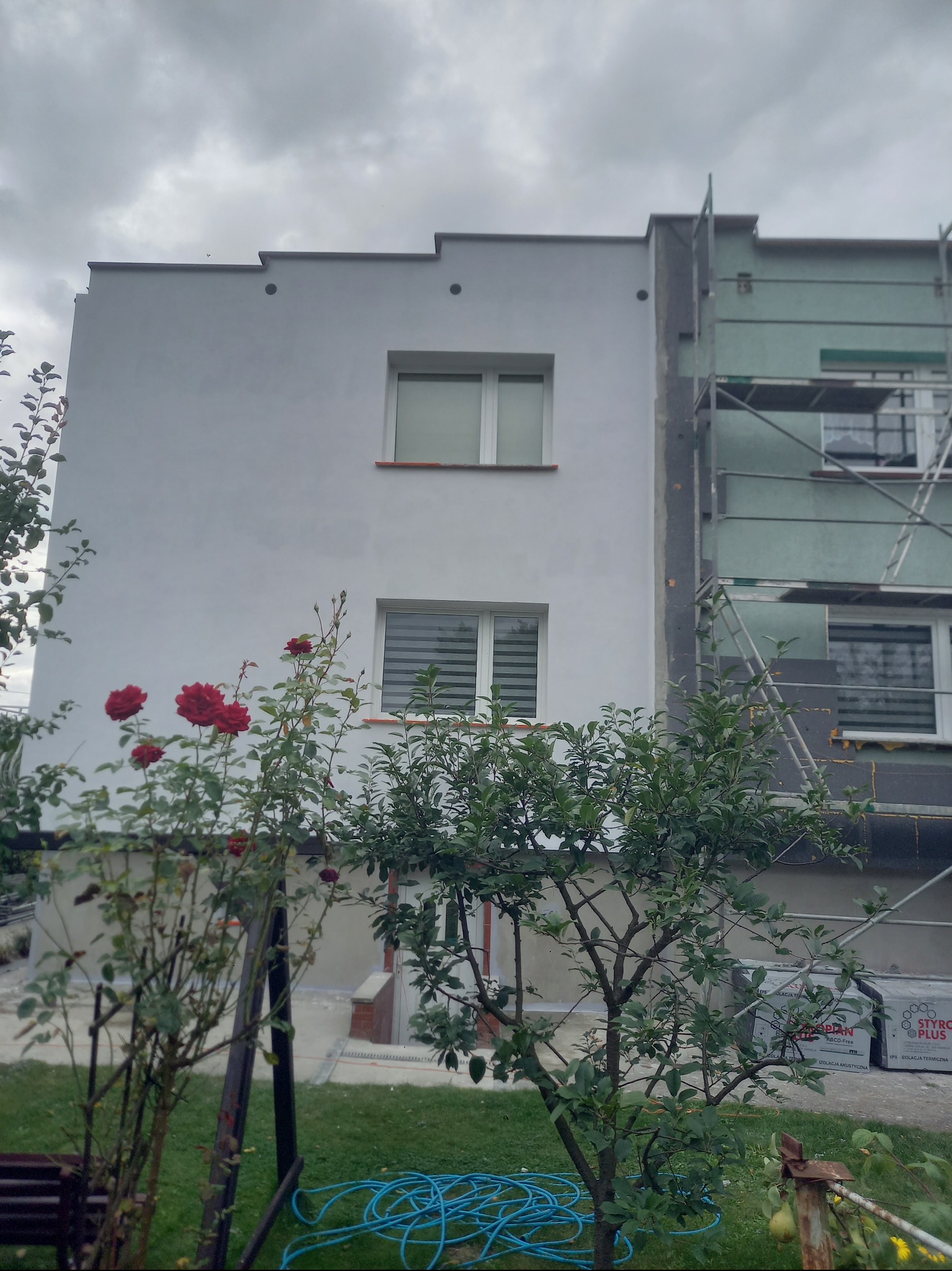 Dom w trakcie ocieplania: jedna ściana pomalowana na biało, druga w trakcie ocieplania styropianem, widoczne rusztowania. W tle pochmurne niebo i roślinność.
