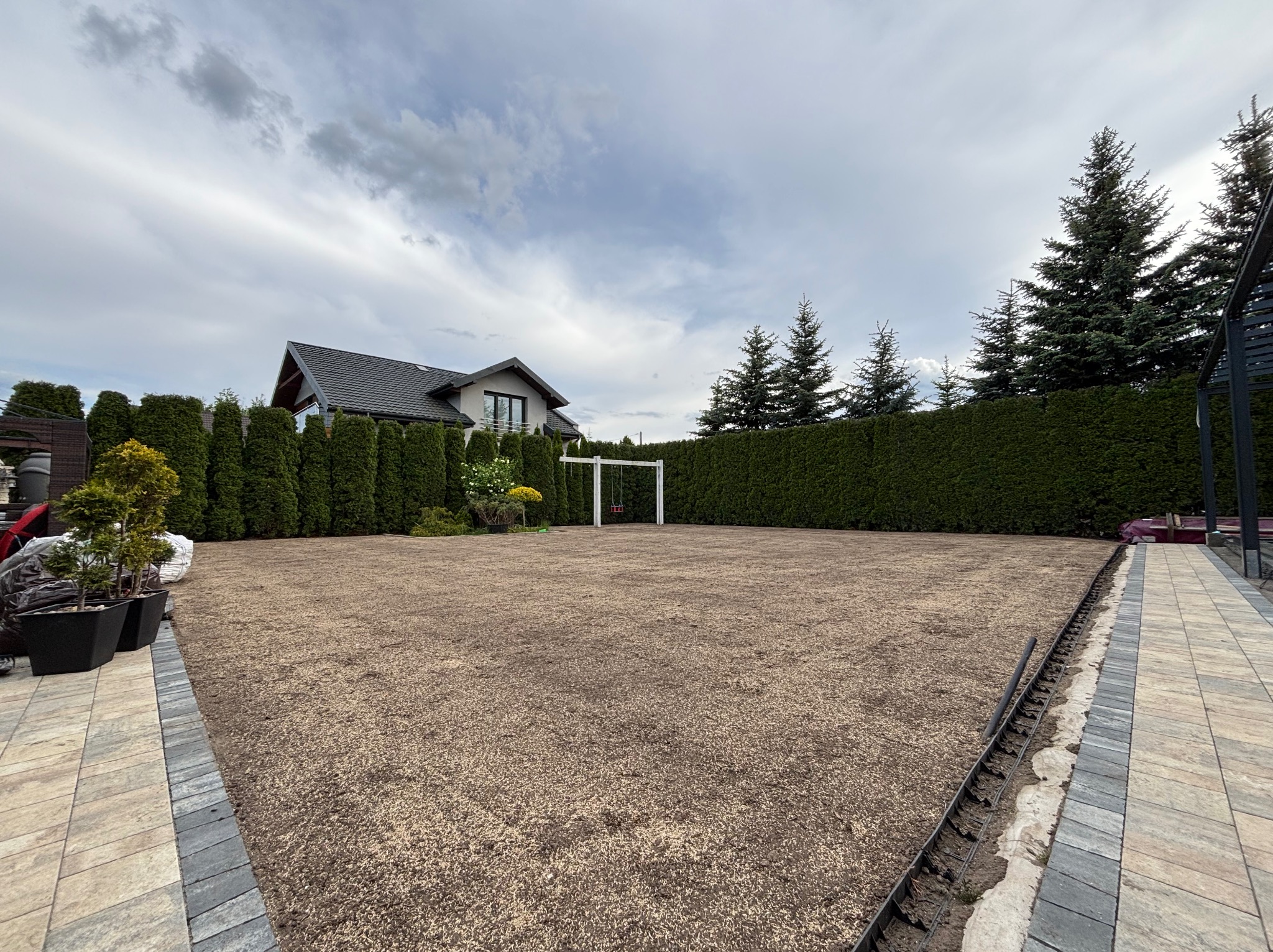 Przygotowany teren pod trawnik z rolki, widoczne obrzeża i podsypka. W tle dom z ogrodem i tujami. Na trawniku huśtawka.