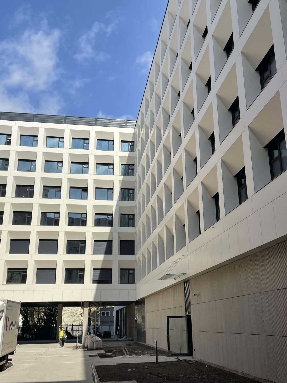 Nowoczesna elewacja budynku z białymi panelami i geometrycznymi oknami, widok z dołu na dziedziniec z pracownikiem w kamizelce odblaskowej i fragmentem samochodu dostawczego.