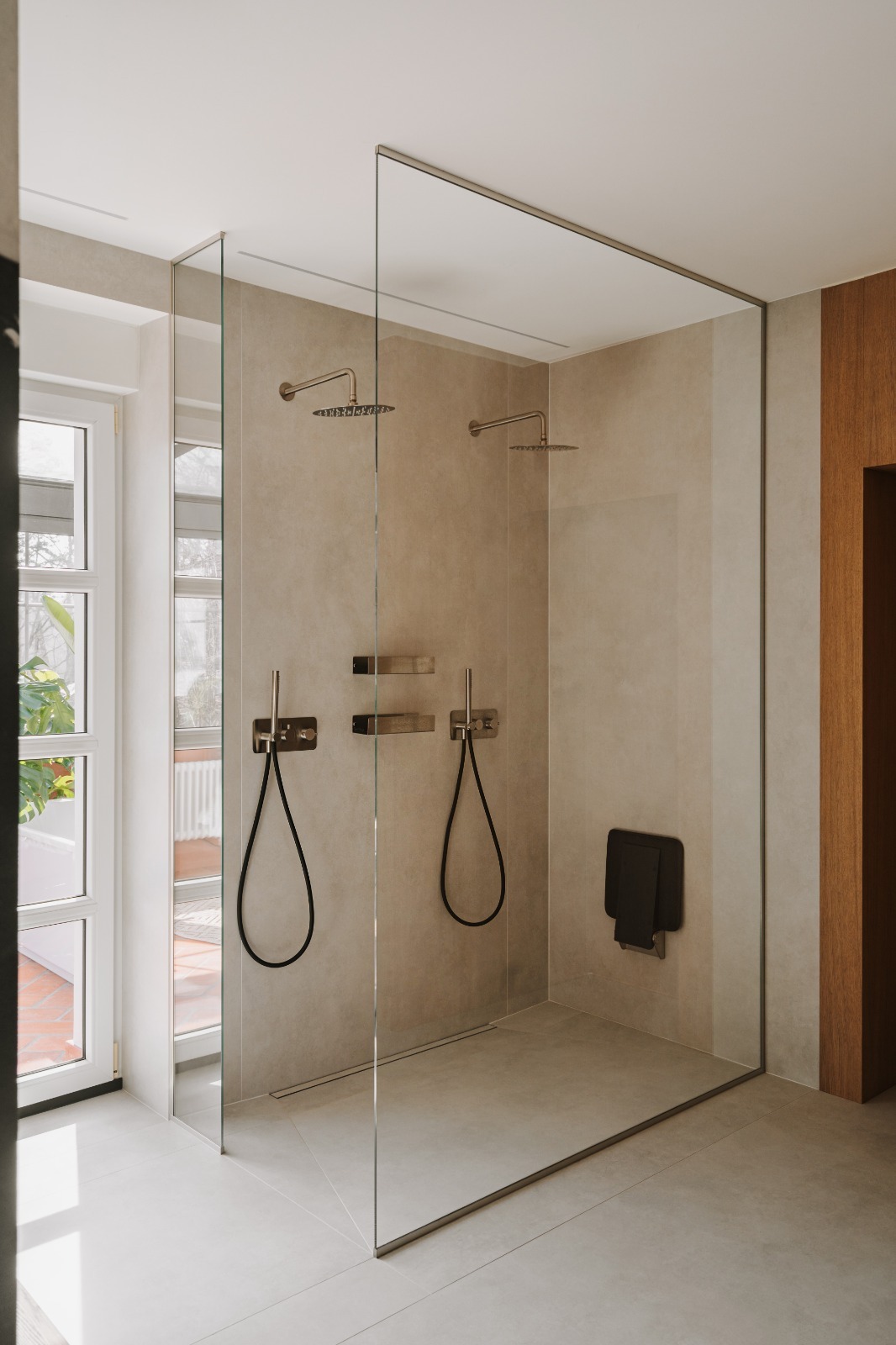 Nowoczesna kabina prysznicowa typu walk-in z dwoma deszczownicami, szklanymi ścianami i minimalistycznym siedziskiem. Beżowe płytki i jasna podłoga tworzą spójną całość.