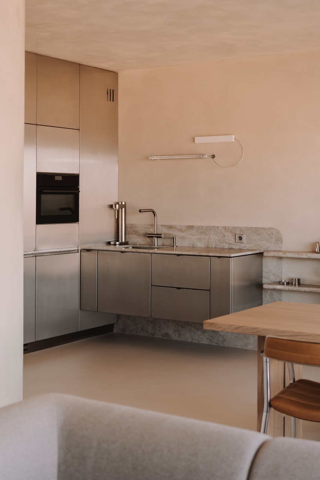 Nowoczesna kuchnia ze stali nierdzewnej i blatem z szarego kamienia. Minimalistyczny design, oświetlenie LED, neutralna kolorystyka ścian. Widoczny fragment stołu i krzesła.