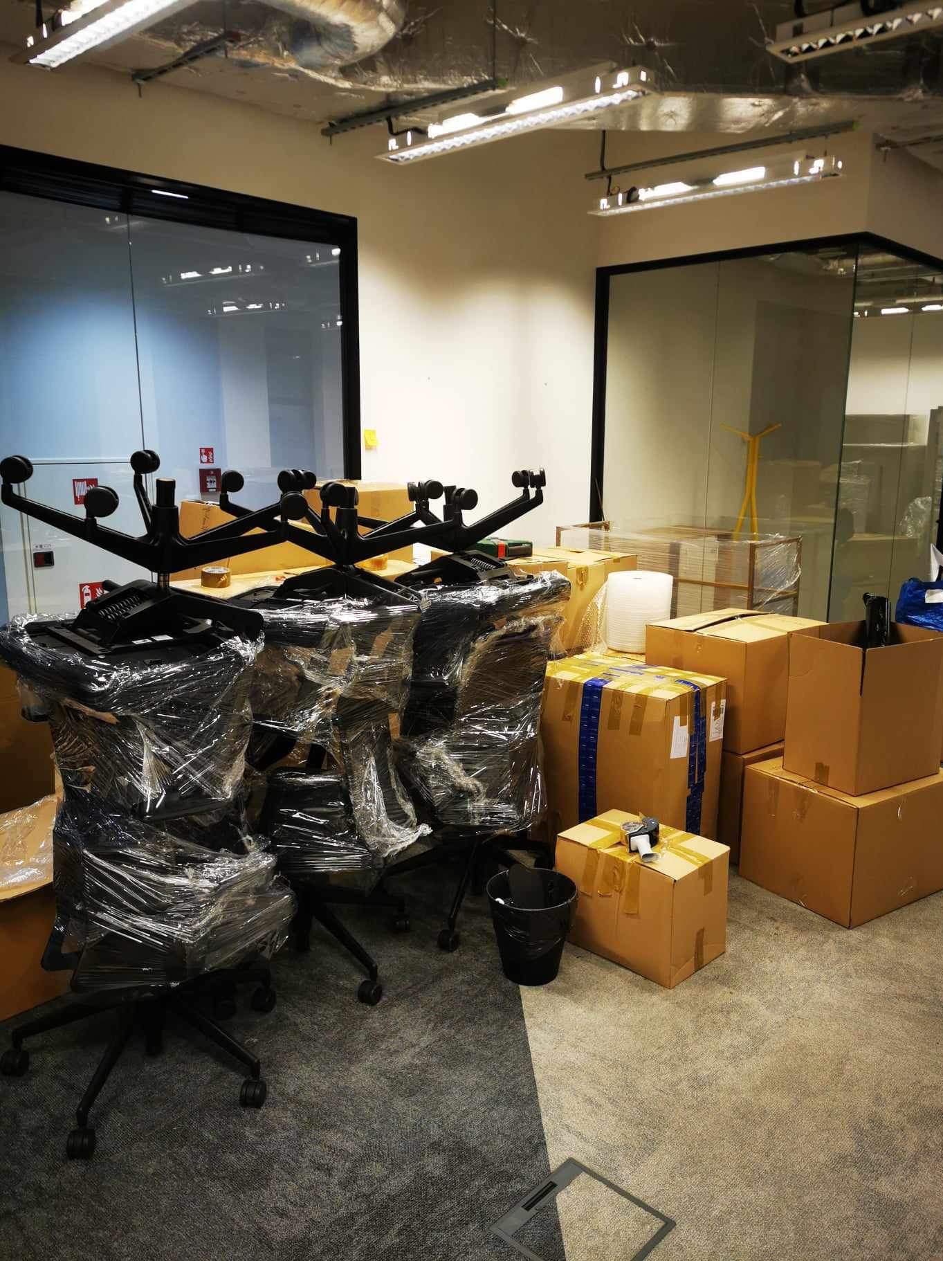 Przygotowane do transportu biurowe krzesła w folii i kartony z taśmą w jasnym biurze z przeszklonymi ścianami. Widoczne elementy instalacji pod sufitem.