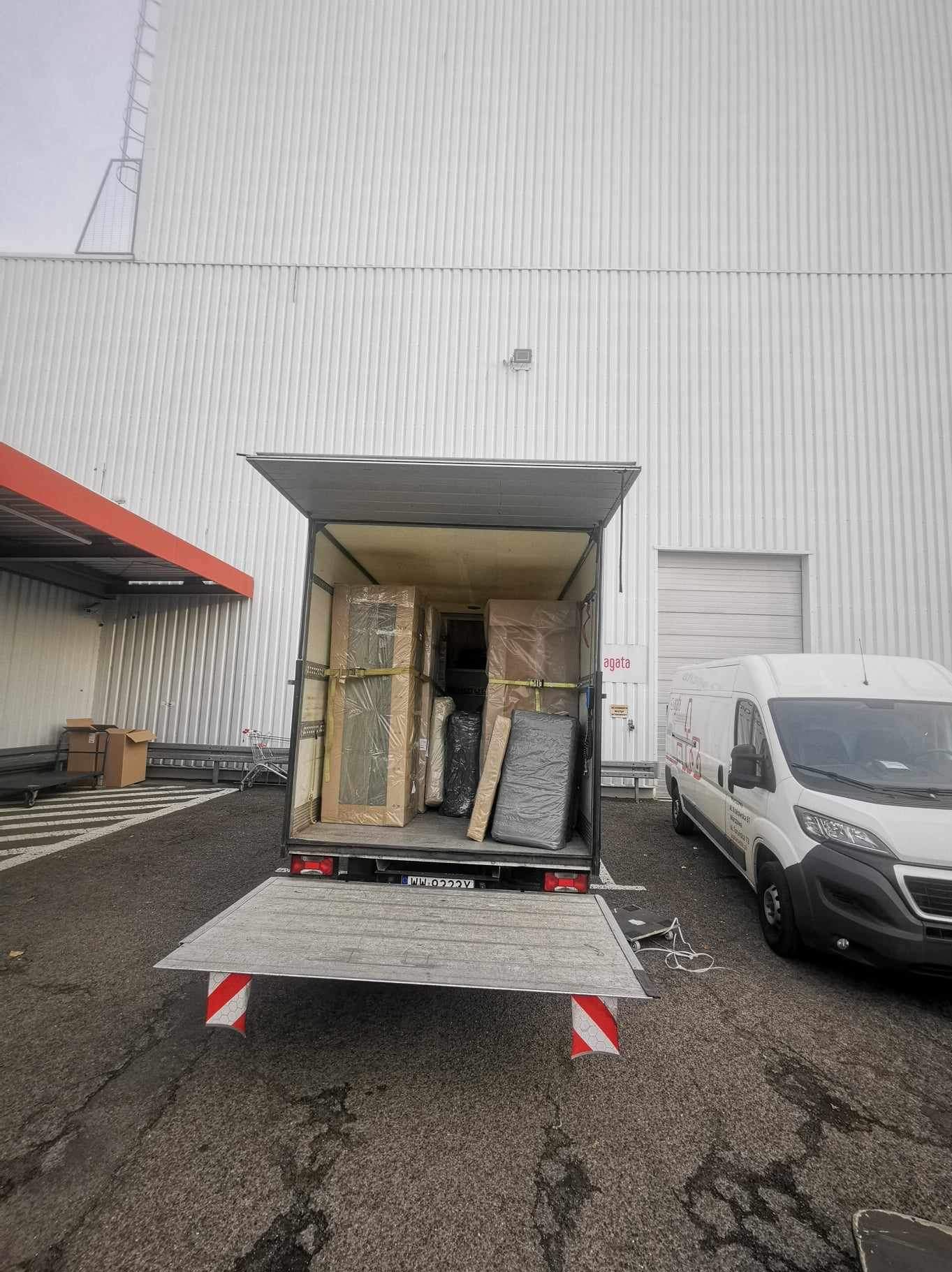 Ciężarówka z otwartą paką załadowana meblami zabezpieczonymi folią i pasami transportowymi, zaparkowana obok białego busa na tle hali magazynowej.