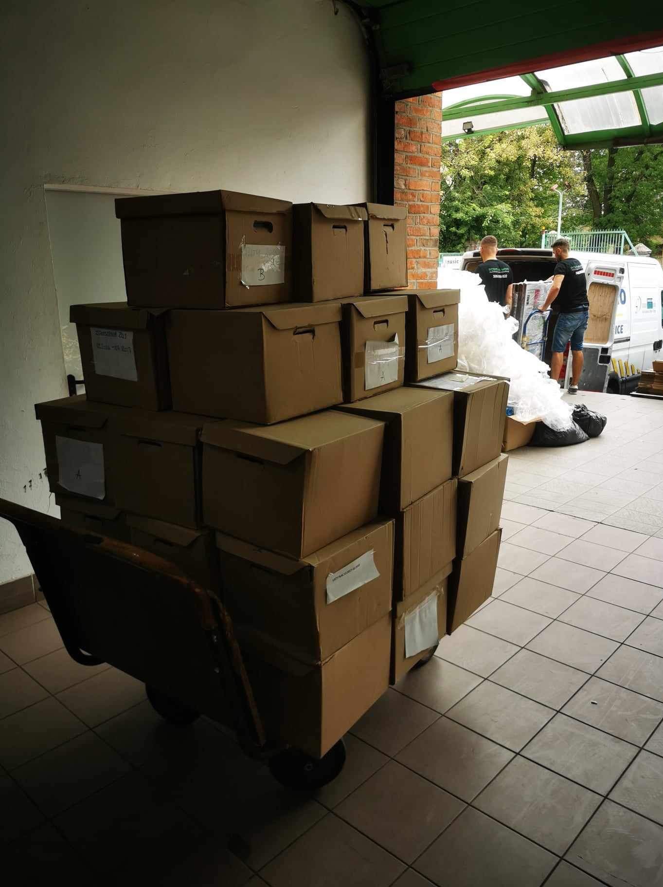Wózek z kartonami, przygotowany do transportu podczas przeprowadzki. W tle pracownicy ładują do busa. Widoczne etykiety na kartonach i ceglana ściana.