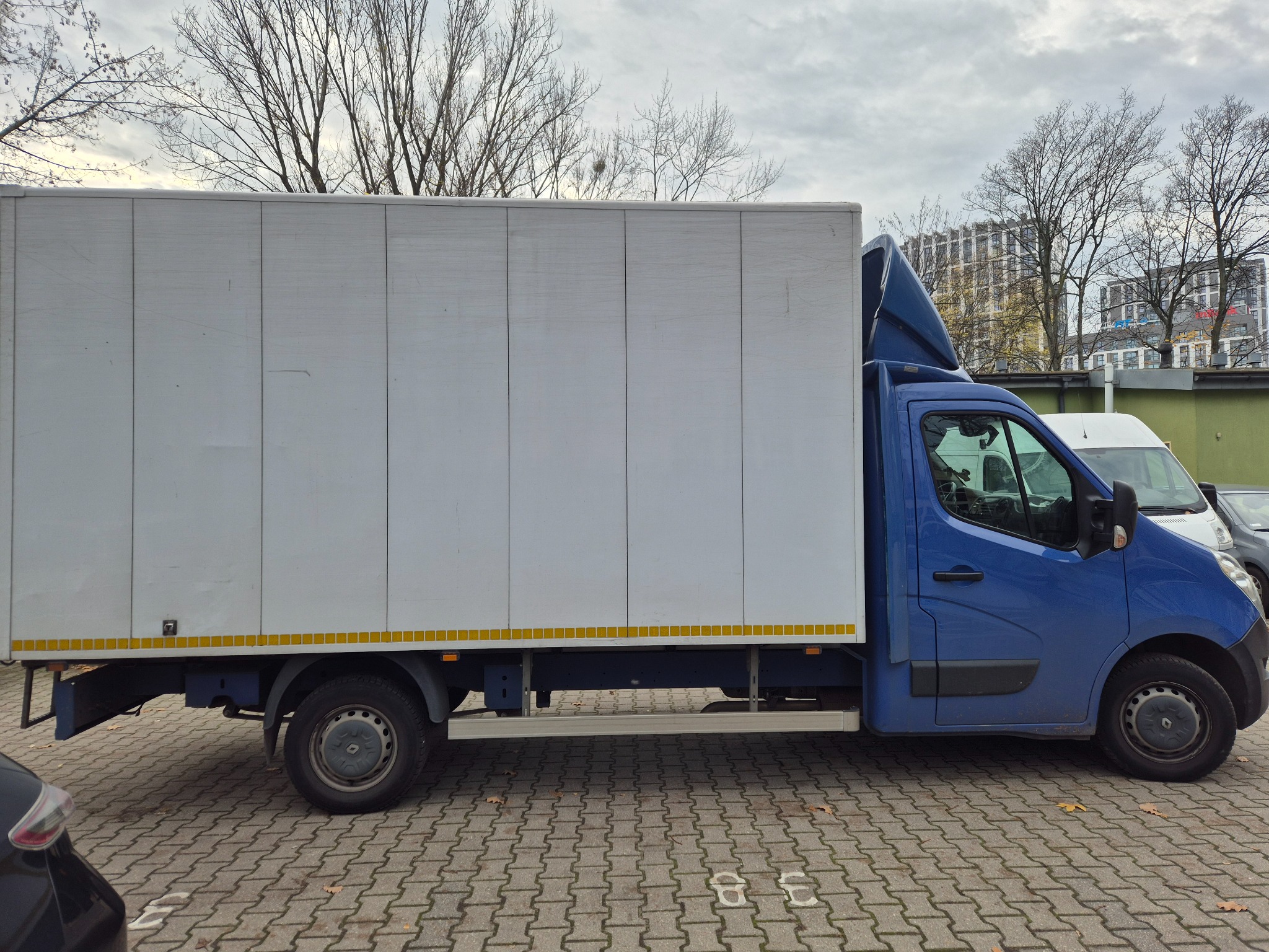 Niebieska furgonetka z białą zabudową kontenerową na parkingu z kostki, widok z boku, drzewa w tle.