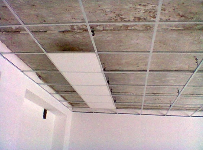 Montaż sufitu podwieszanego: widoczne metalowe profile i częściowo zamontowane białe panele. Surowy betonowy strop w tle. Ujęcie z dołu, z narożnikiem ściany.