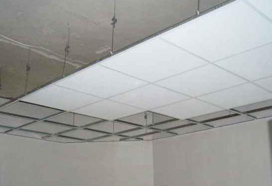 Kwadratowy sufit podwieszany z widoczną metalową konstrukcją i białymi panelami, zawieszony na linkach w jasnym pomieszczeniu. Widok z dołu.