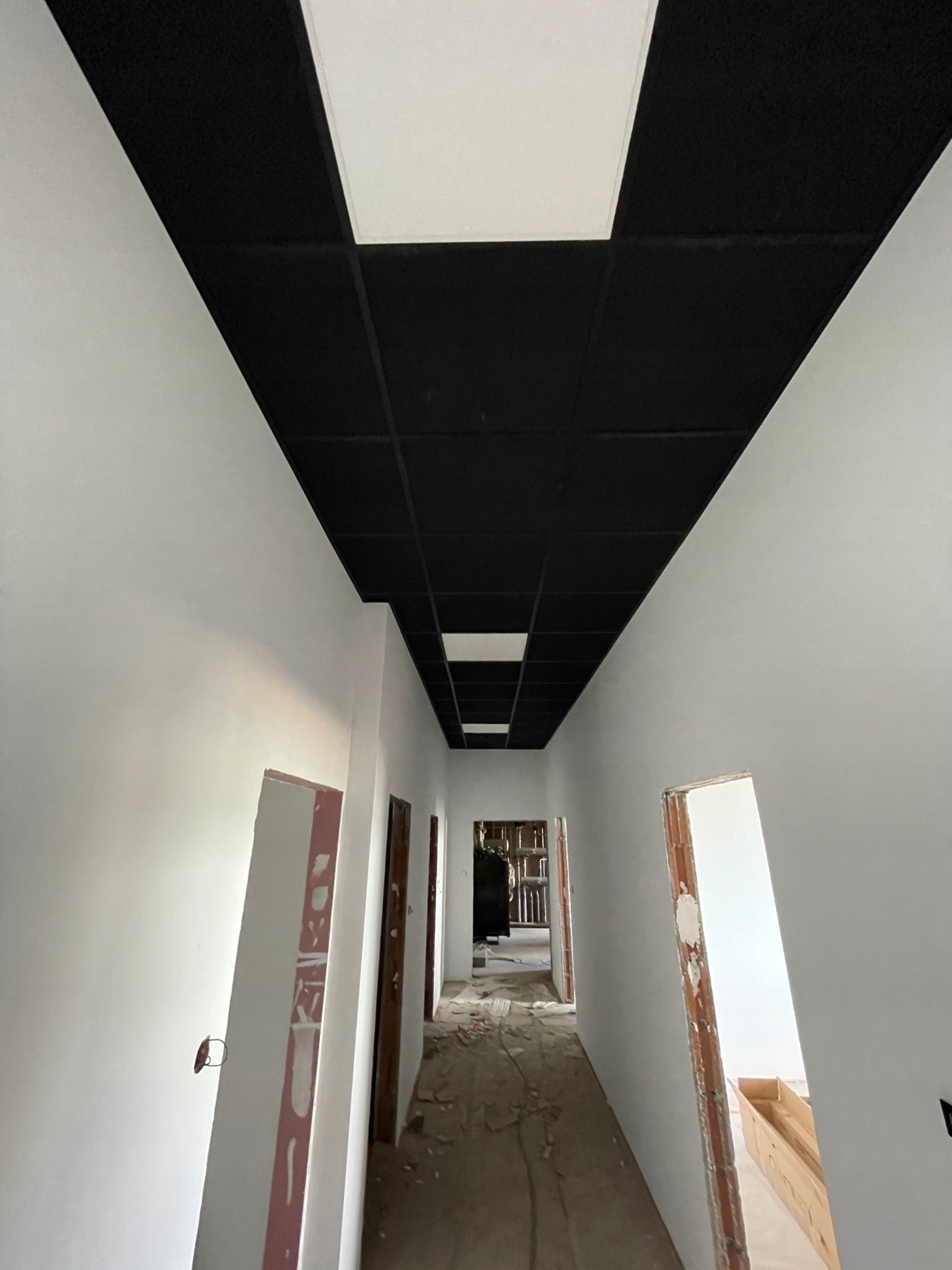 Korytarz w trakcie wykańczania: białe ściany, czarny sufit kasetonowy z białymi elementami. Widoczne niedokończone ościeżnice i podłoga w trakcie prac.