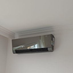 BG KLIMATYZACJA Bogdan Głowacz - Szary klimatyzator ścienny marki Versu, zamontowany w jasnym pomieszczeniu z dekoracyjną listwą sufitową. Widoczne odbicie okna w obudowie urządzenia.