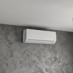 BG KLIMATYZACJA Bogdan Głowacz - Biały klimatyzator ścienny Haier na szarej ścianie o fakturze betonu, zamontowany pod białym sufitem. Widok z dołu, oświetlenie dzienne.