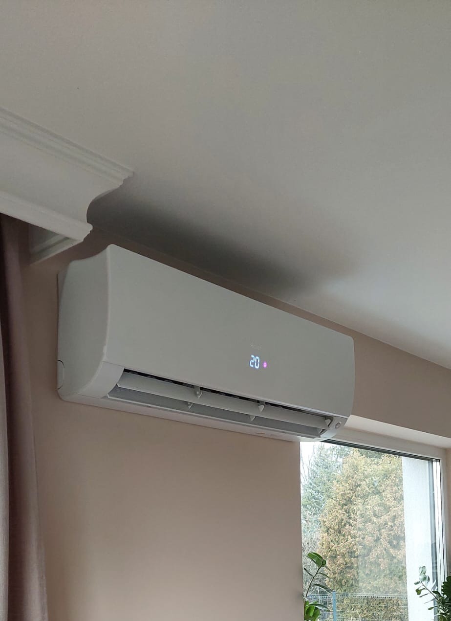 Biały klimatyzator ścienny Haier z wyświetlaczem temperatury 20 stopni, zamontowany w jasnym wnętrzu z oknem i dekoracyjną listwą sufitową.