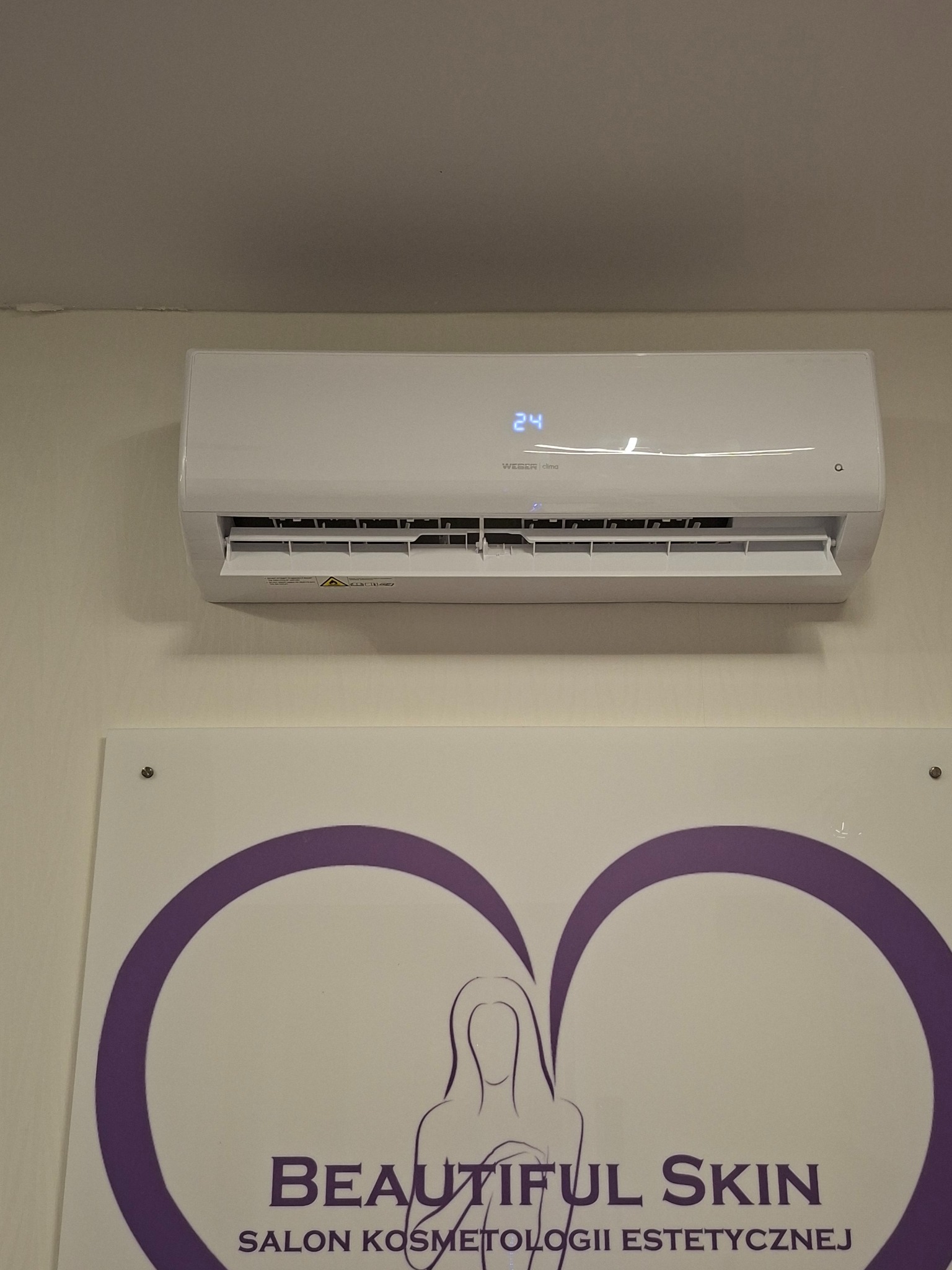 Biała klimatyzacja WEBER clima zamontowana nad logo salonu kosmetycznego 'Beautiful Skin'. Widoczny wyświetlacz z temperaturą 24 stopnie.
