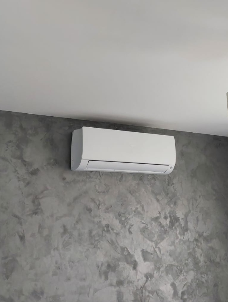 Biały klimatyzator ścienny Haier na szarej ścianie o fakturze betonu, zamontowany pod białym sufitem. Widok z dołu, oświetlenie dzienne.