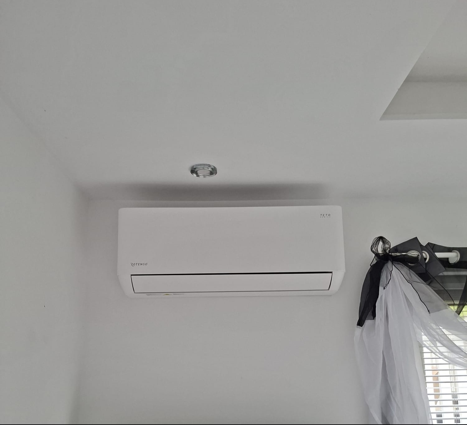 Biały klimatyzator Rotenso Teta zamontowany na ścianie w jasnym pomieszczeniu, widoczny fragment okna z firanką. Minimalistyczny design.
