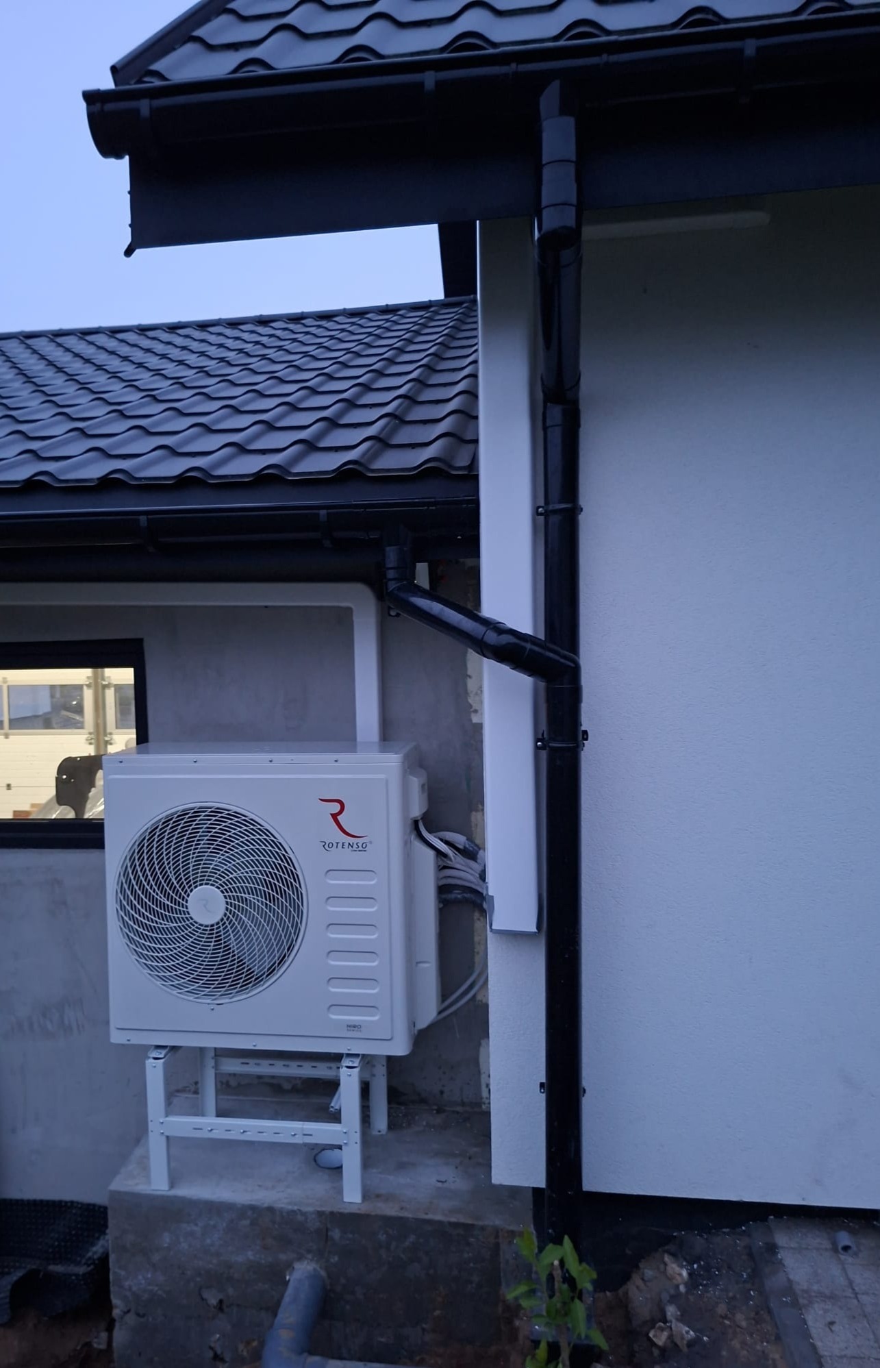 Biała jednostka zewnętrzna klimatyzacji Rotenso zamontowana na metalowym stelażu przy szarej ścianie budynku z czarną rynną i dachem z dachówki.