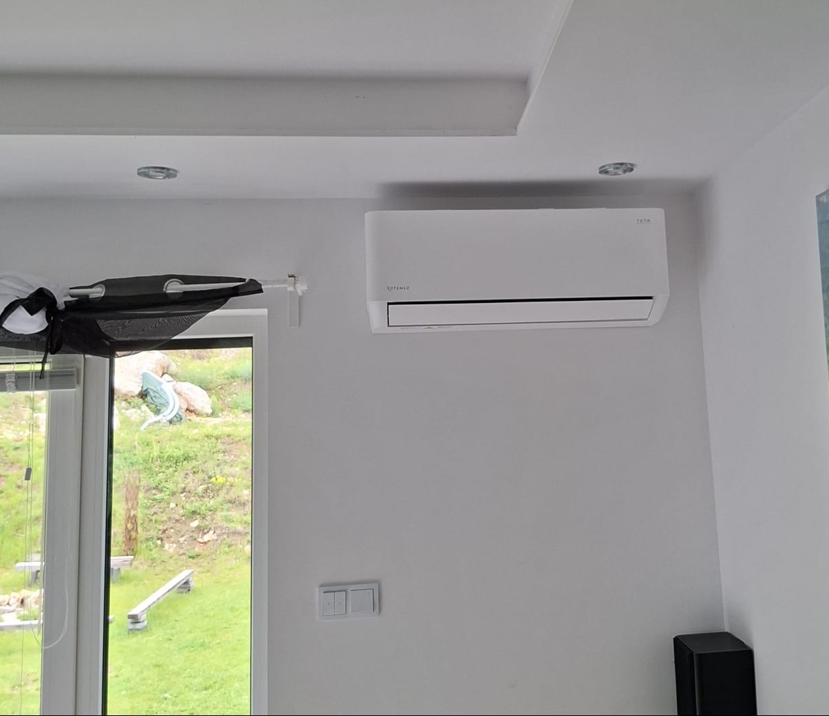 Biały klimatyzator ROTENSO zamontowany na ścianie w jasnym wnętrzu, widok z okna na zewnątrz, minimalistyczny design, nowoczesne oświetlenie sufitowe.