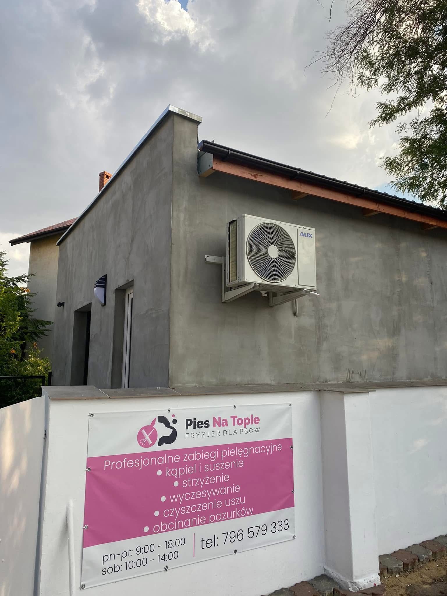 Szary budynek z klimatyzatorem AUX i szyldem salonu fryzjerskiego dla psów 'Pies Na Topie'. Widoczne zabiegi pielęgnacyjne i numer telefonu.