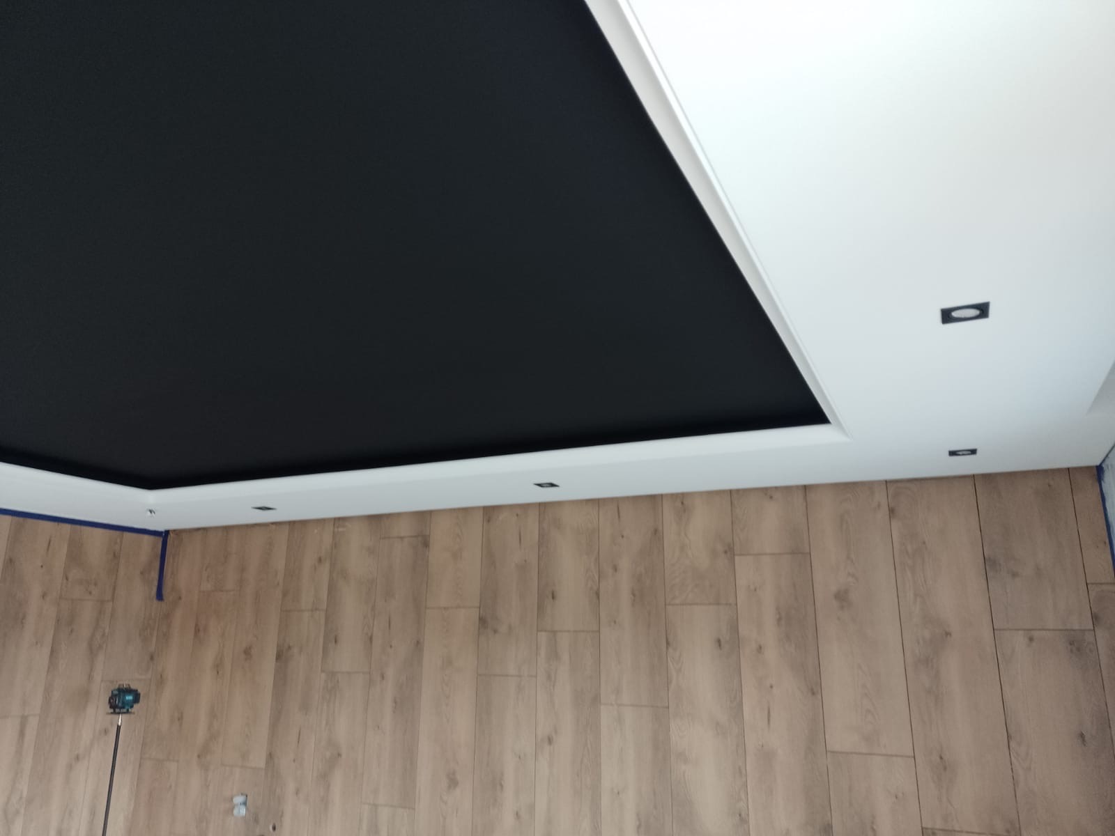 Nowoczesny sufit podwieszany z czarnym panelem i oświetleniem punktowym, wykończony białą listwą. Parkiet na podłodze. Widoczny laser krzyżowy.