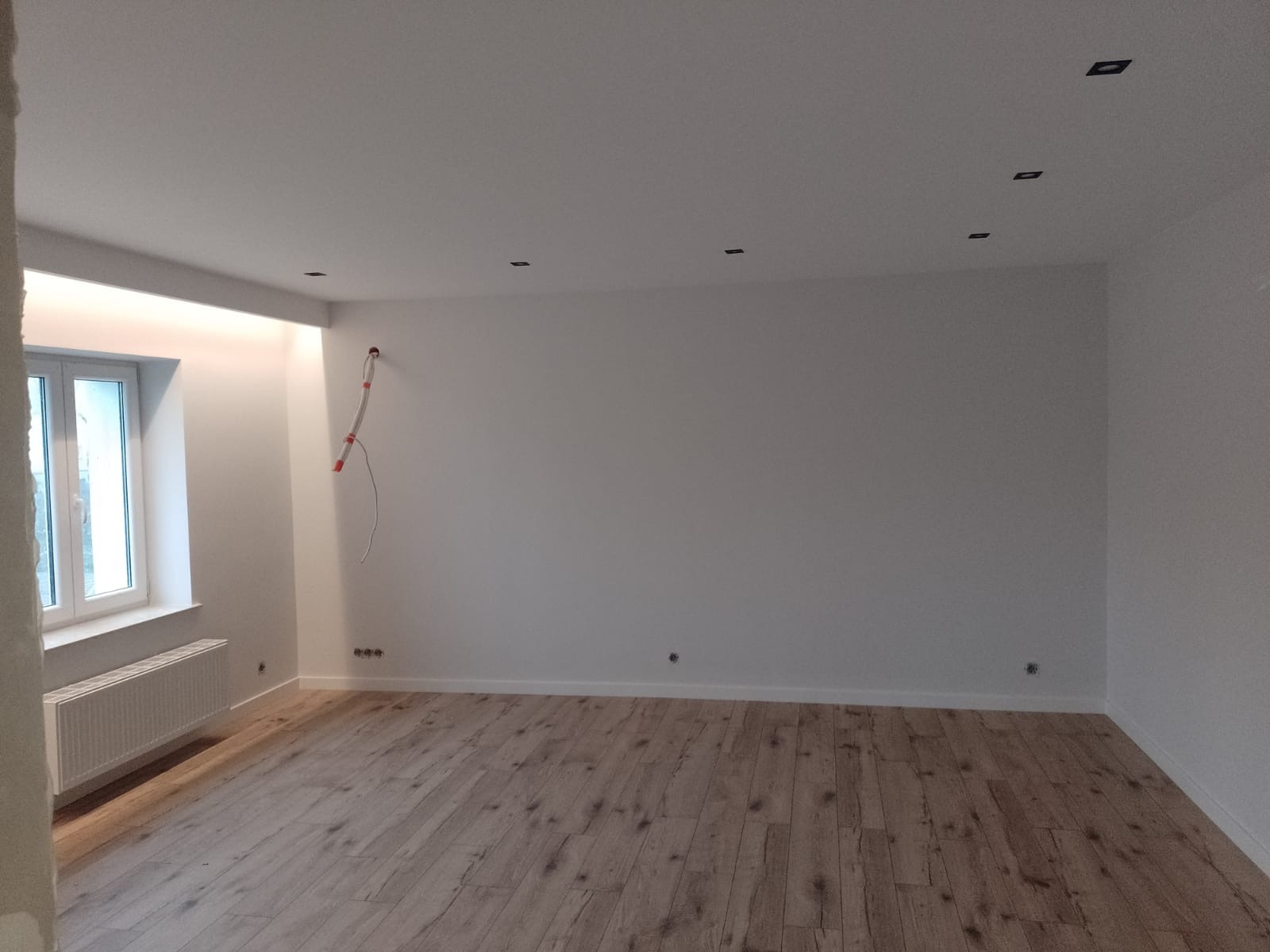 Wnętrze po remoncie w Mazurowicach: świeżo pomalowane ściany na biało, drewniana podłoga, okno z grzejnikiem i przygotowane punkty świetlne w suficie.