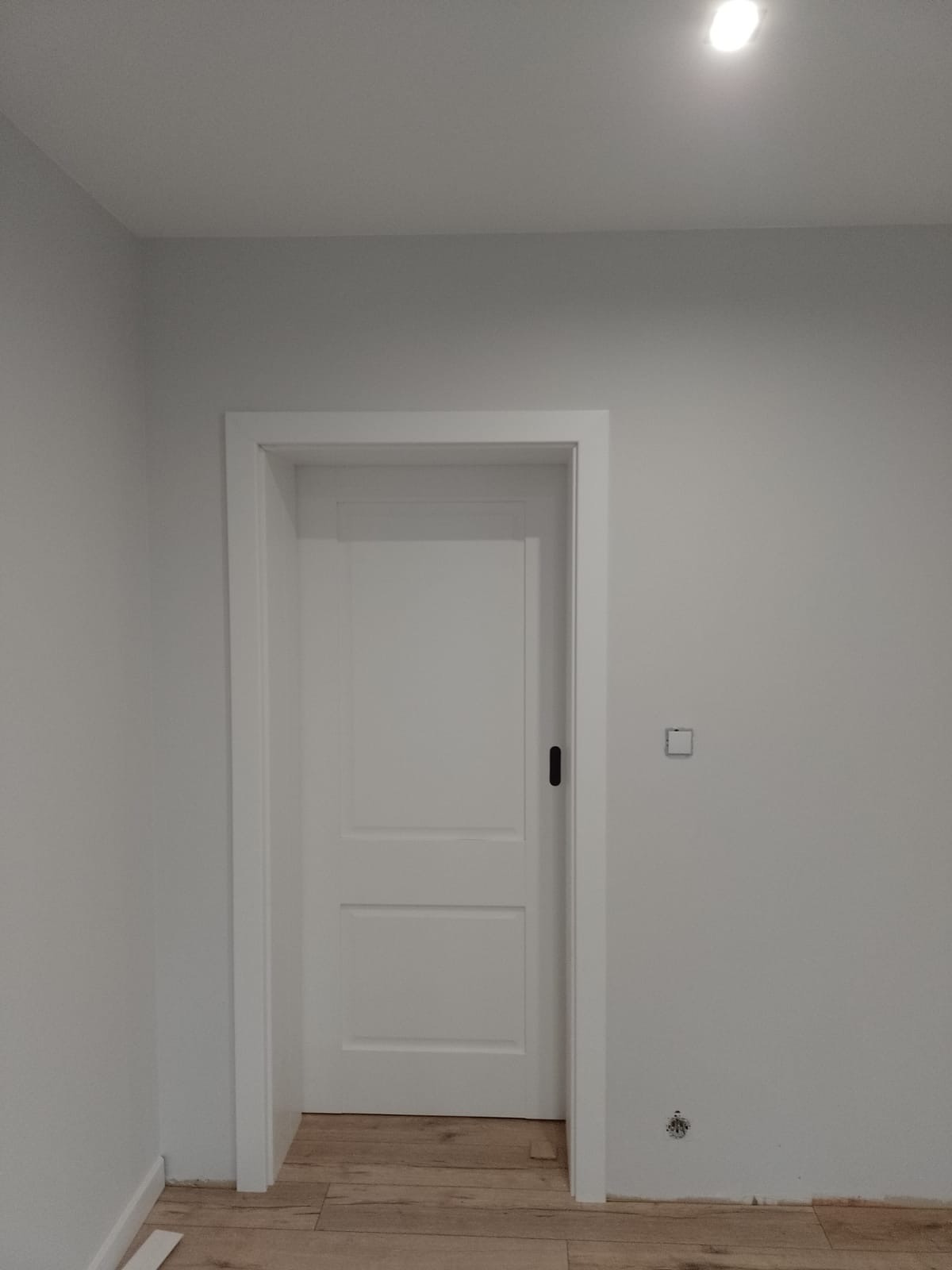 Białe, dwuskrzydłowe drzwi wewnętrzne z czarnym uchwytem, osadzone w białej futrynie na tle szarej ściany i drewnianej podłogi. Widoczny fragment instalacji elektrycznej.