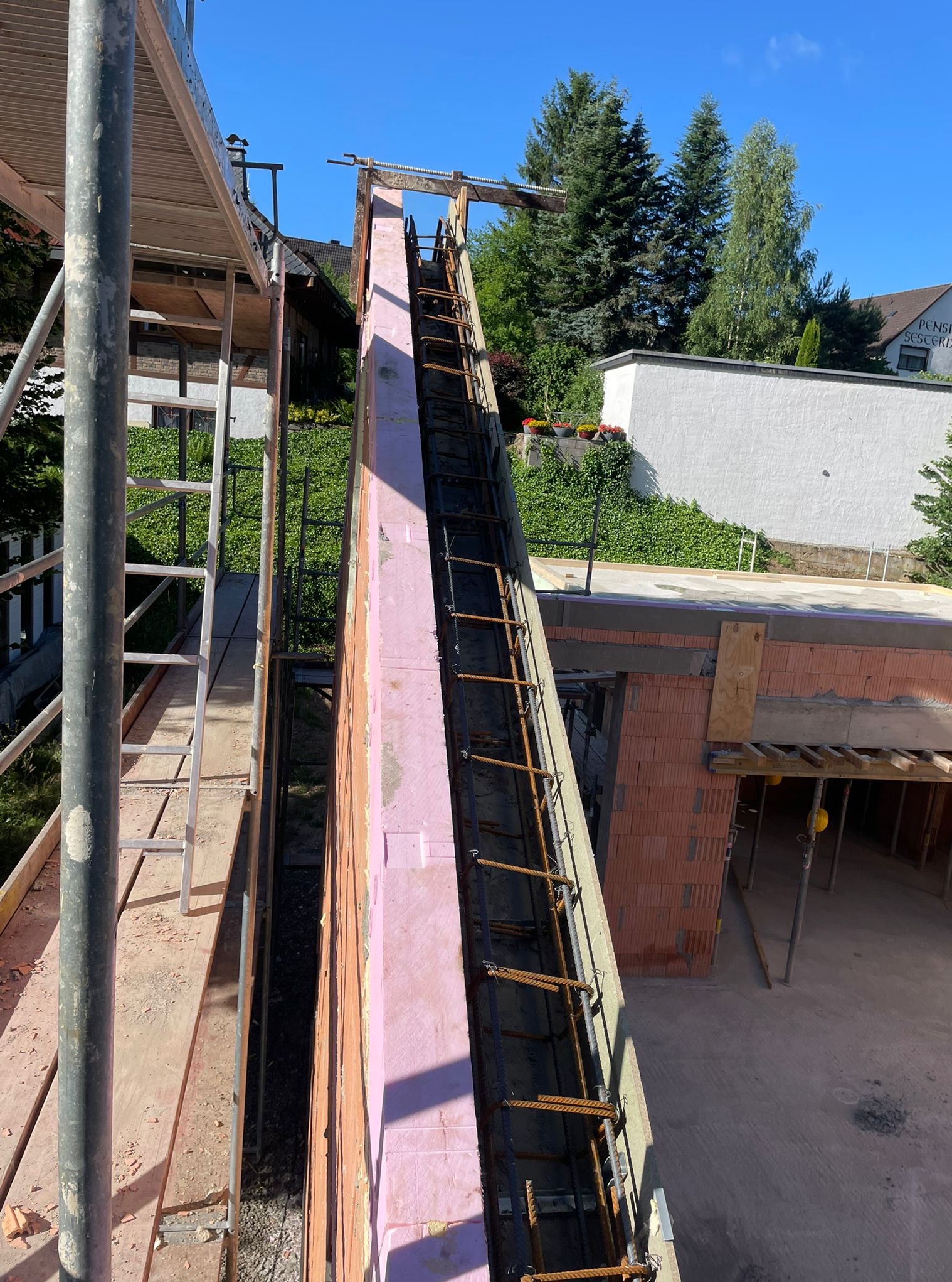 Wysoka ściana z różowym ociepleniem i zbrojeniem, widok z rusztowania na budowę domu z cegły, w tle zieleń i budynki.