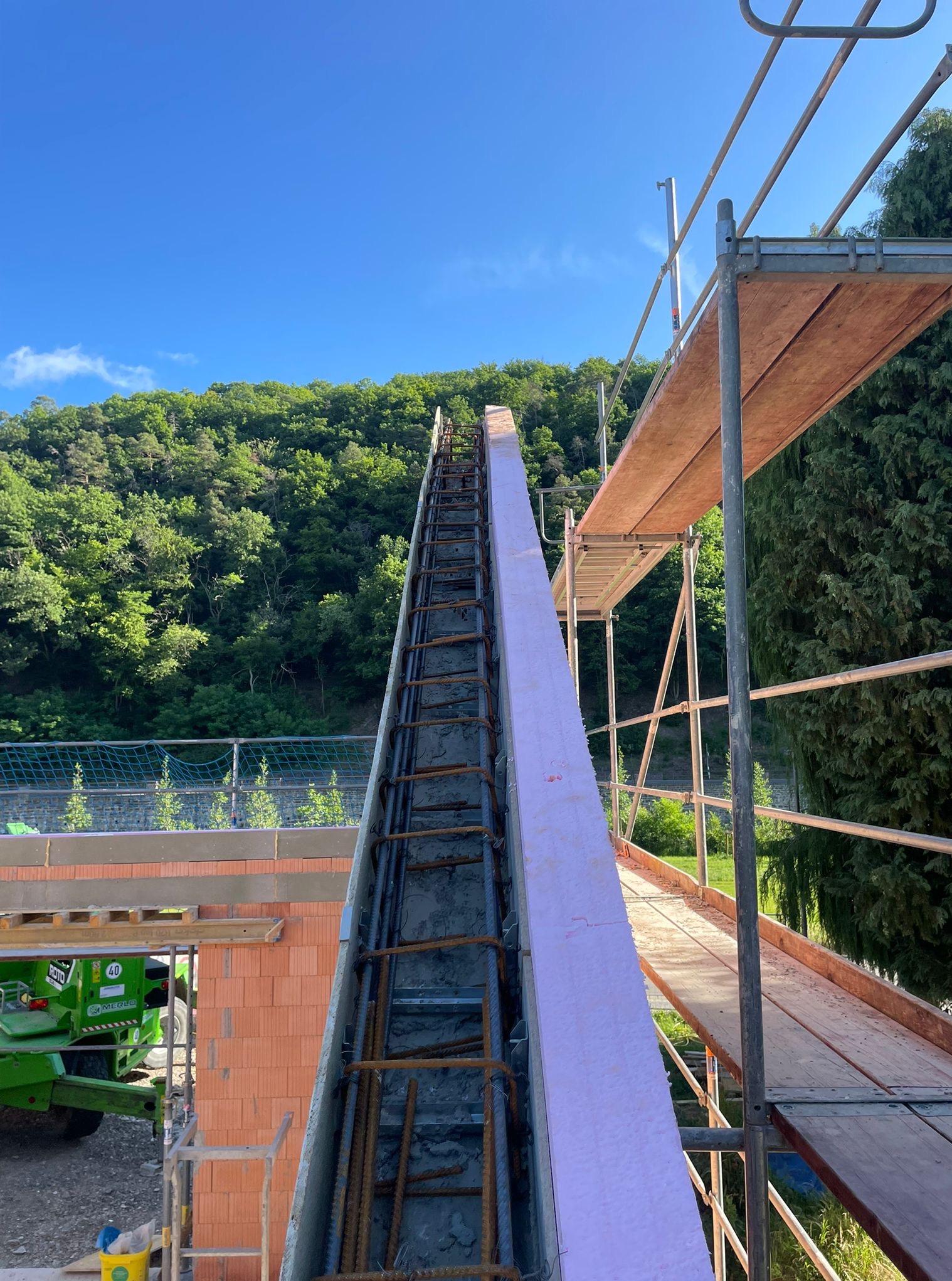 Wąski szalunek zbrojony, wypełniony betonem, z różową izolacją, ustawiony pionowo na tle zieleni i błękitnego nieba. Widoczne rusztowanie i fragment ściany z cegieł.