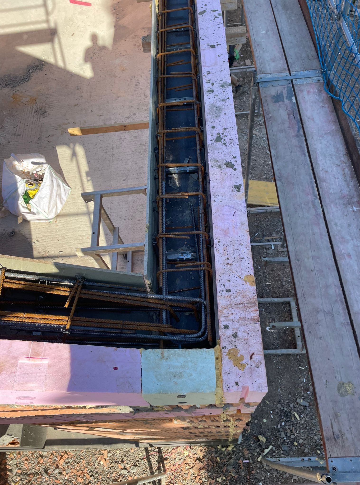 Zbrojenie pionowe w szalunku z termoizolacją, widok z góry. Element konstrukcyjny w trakcie budowy, z rusztowaniem w tle. Detal budowlany.