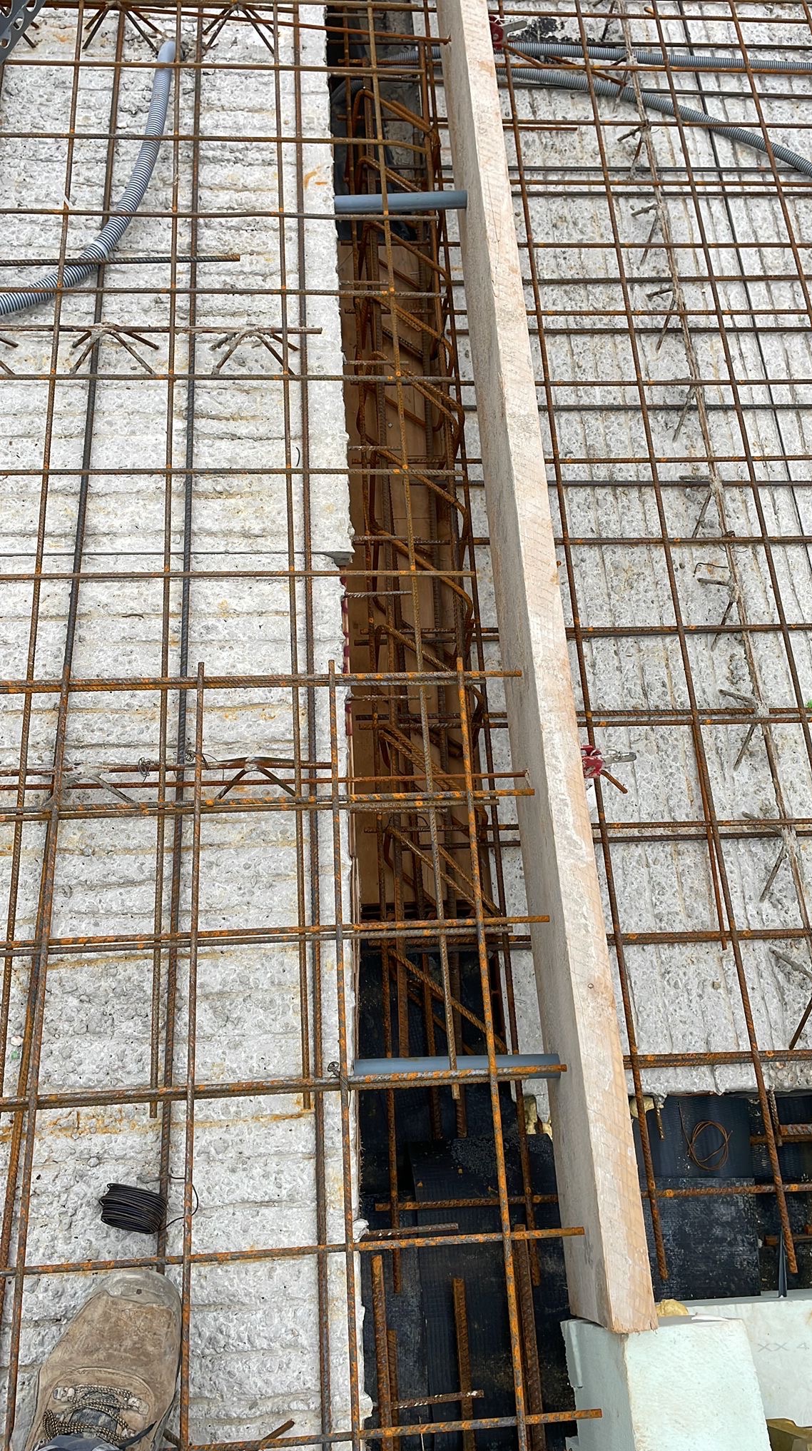 Zbrojenie fundamentu z drewnianym szalunkiem i instalacją, widok z góry. Siatka z prętów zbrojeniowych, fragment buta roboczego w lewym dolnym rogu.