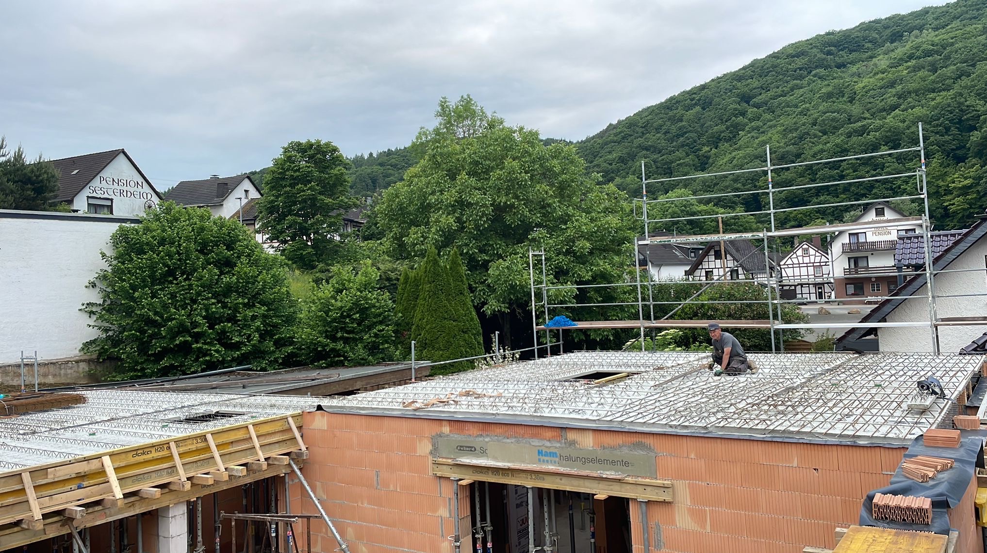 Budowa domu z czerwonej cegły: pracownik montuje zbrojenie na stropie. Widoczne szalunki, rusztowania i zielone wzgórza w tle. Ujęcie z góry, w dzień.