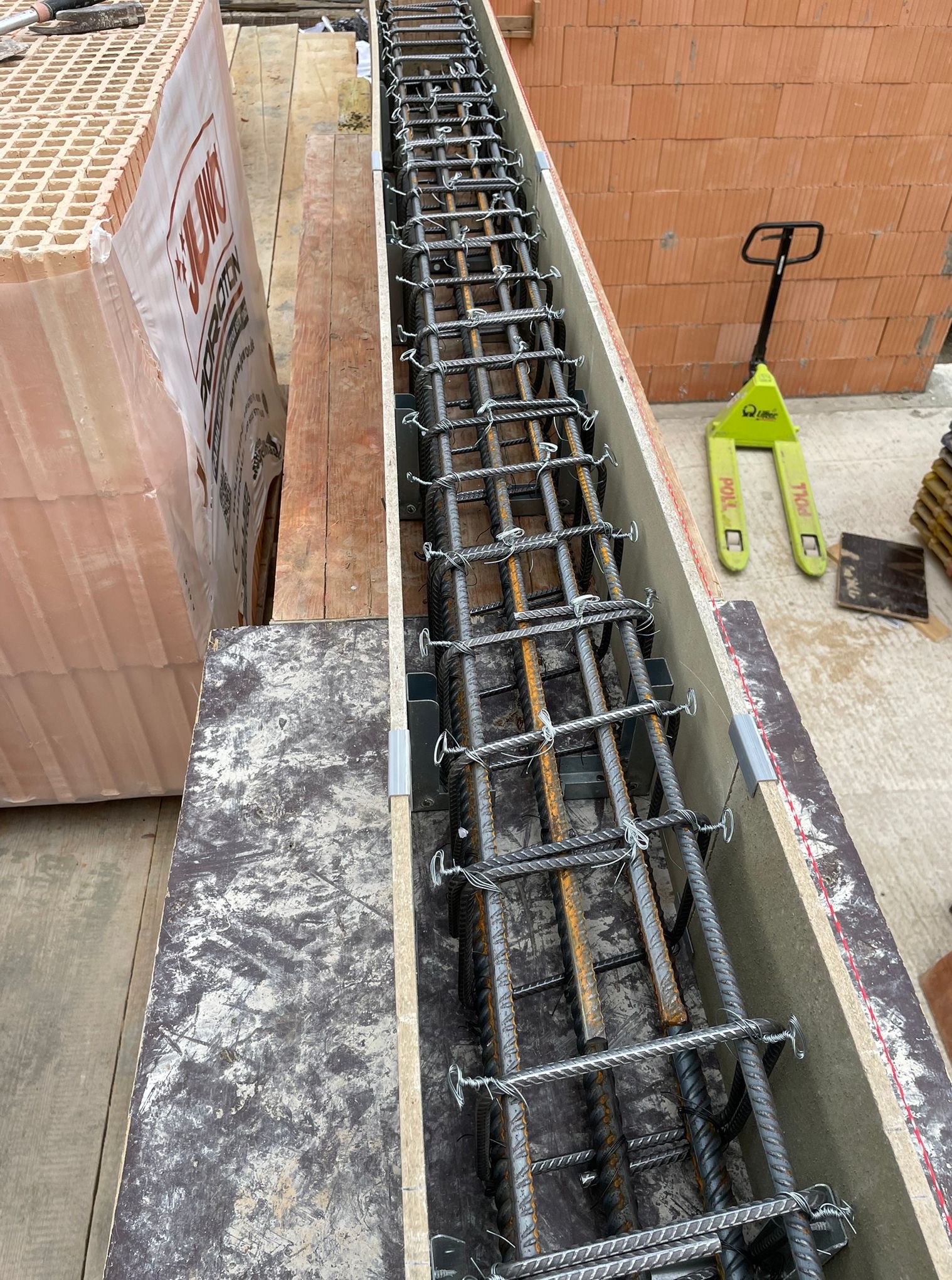 Zbrojenie w szalunku, widok z góry. Stalowe pręty połączone drutem, przygotowane do zalania betonem. W tle bloczki ceramiczne i wózek paletowy.