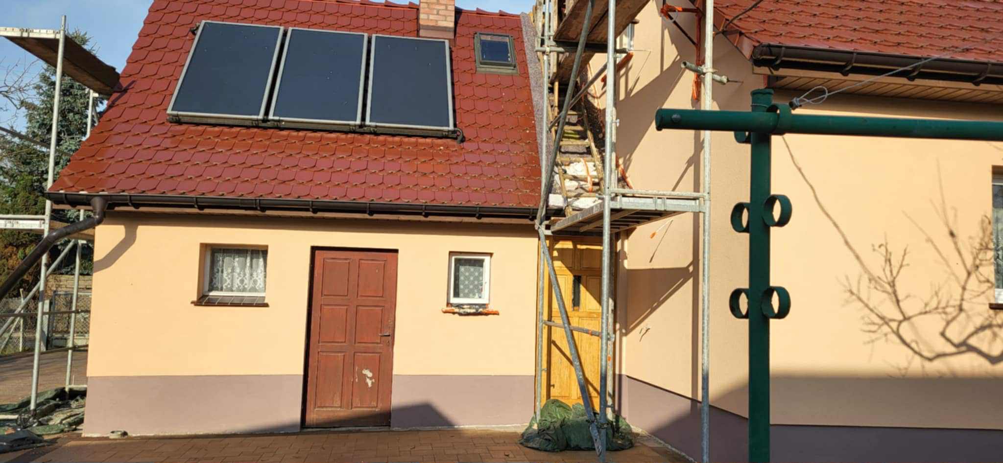 Dwukondygnacyjny dom z panelami słonecznymi na czerwonym dachu, częściowo zasłonięty rusztowaniem. Ściany w odcieniach beżu i brązu. Widoczne okna i drzwi.