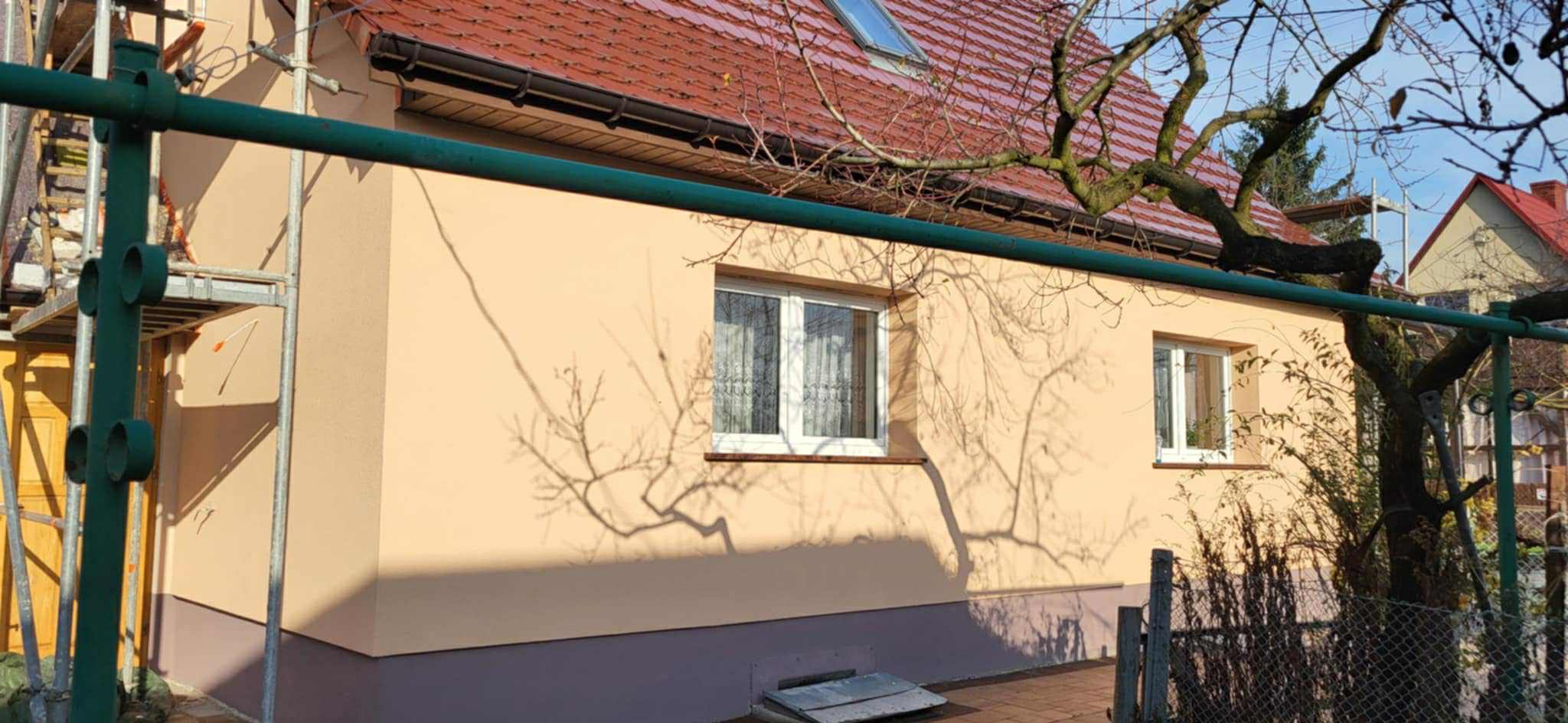 Ocieplona elewacja domu jednorodzinnego w beżowym odcieniu, z widocznymi oknami i cieniem drzewa. Fragment rusztowania po lewej stronie. Dach z czerwonej dachówki.