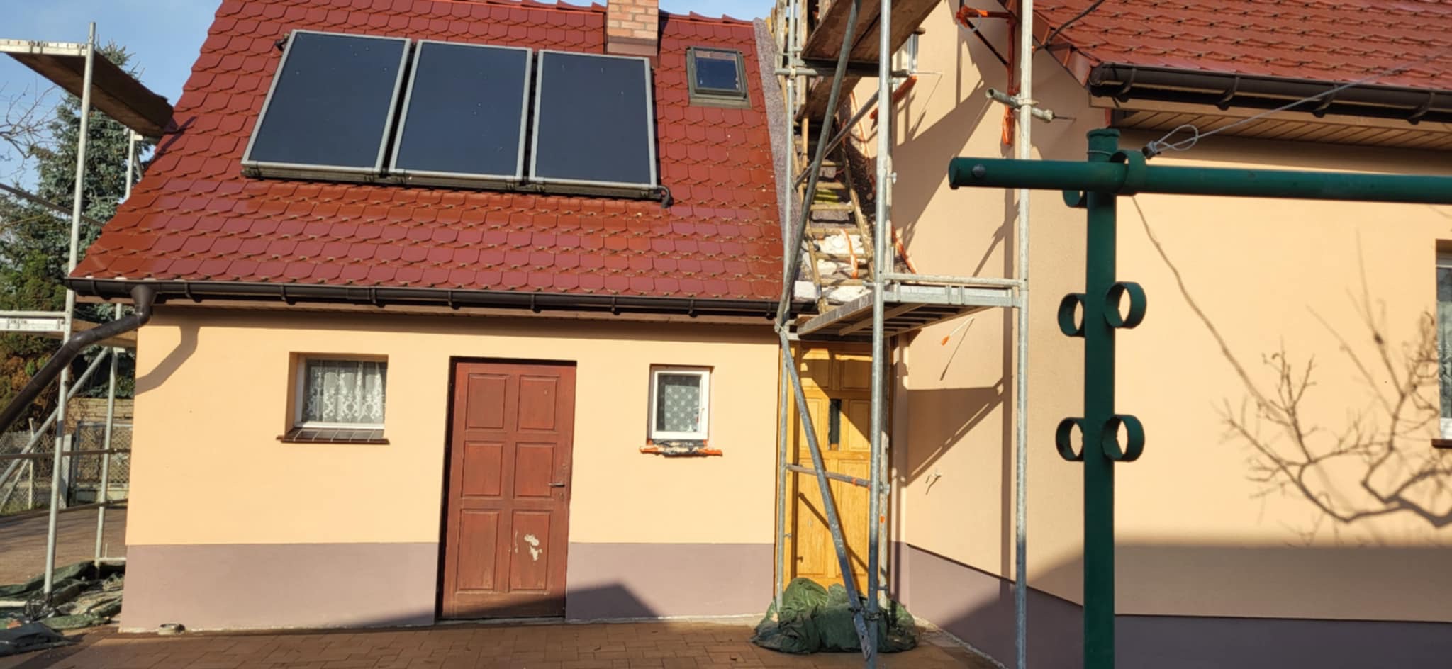 Dom jednorodzinny z panelami słonecznymi na czerwonym dachu, częściowo zasłonięty rusztowaniem. Ściany w odcieniach beżu i brązu. Widoczne okna i drewniane drzwi.