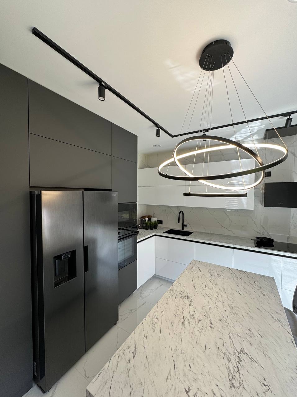 Nowoczesna kuchnia z szarymi szafkami, marmurowym blatem i designerską lampą pierścieniową. Oświetlenie szynowe podkreśla minimalistyczny charakter wnętrza.