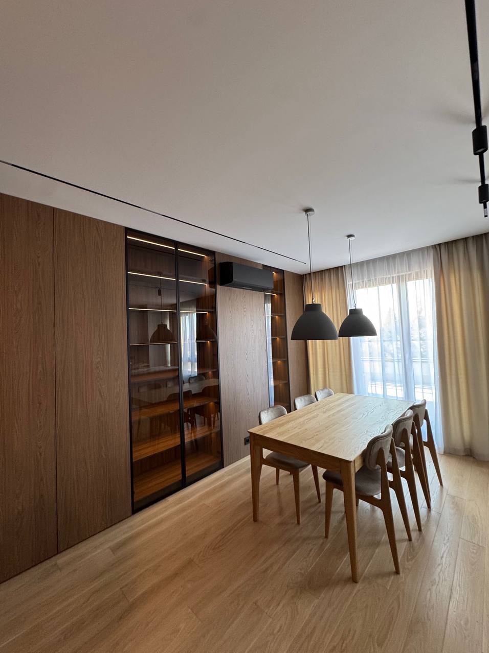 Nowoczesne wnętrze jadalni z drewnianą zabudową ścienną, przeszkloną witryną z oświetleniem LED i podwieszanym klimatyzatorem. Dwa czarne klosze lamp nad stołem.