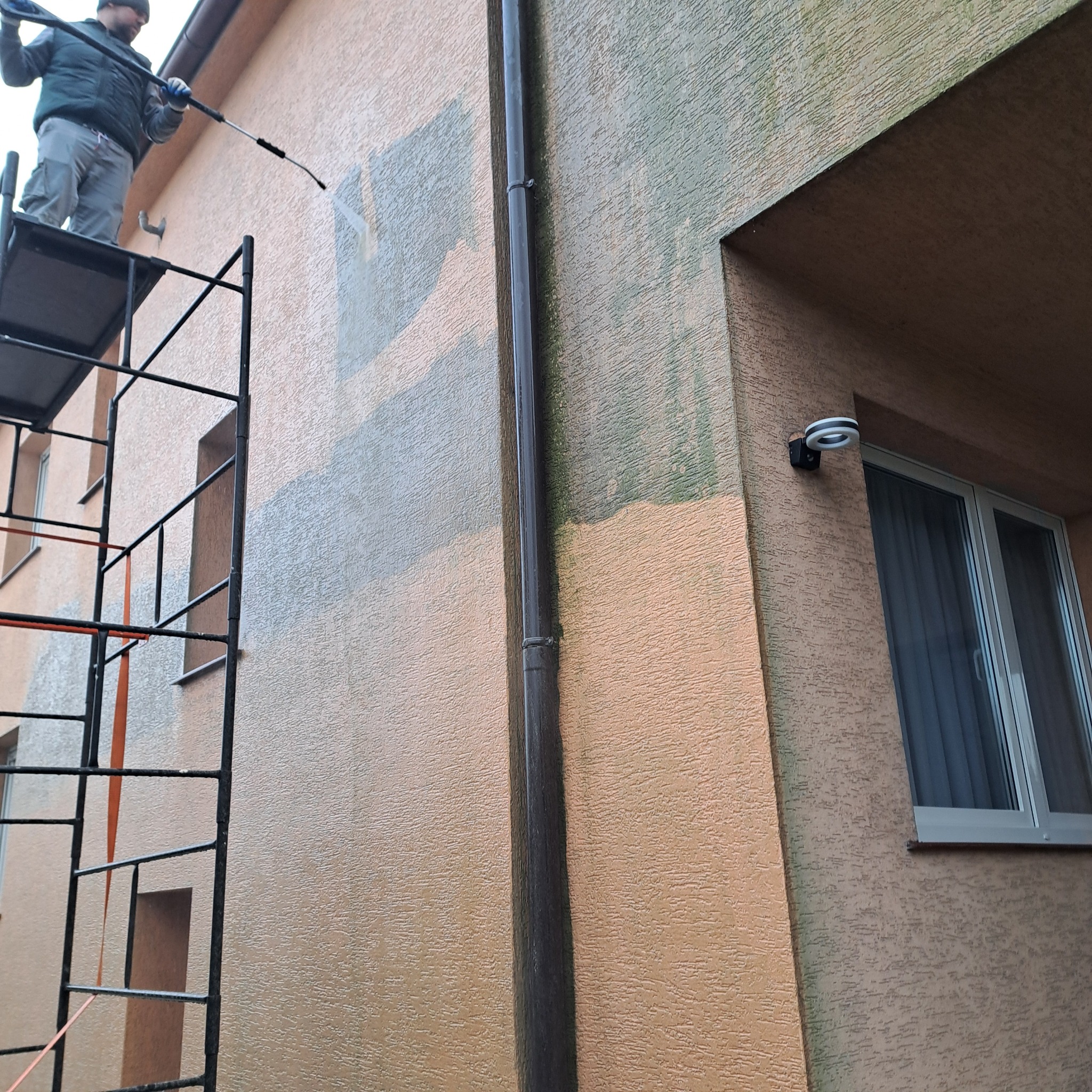 Czyszczenie elewacji budynku z rusztowania za pomocą myjki ciśnieniowej. Widoczne fragmenty umytej ściany, rynna i okno. Praca na wysokościach.