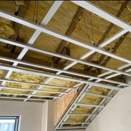 KOWBUD Konrad Kowalski - Sufit podwieszany z izolacją z wełny mineralnej, widoczne profile metalowe i drewniana konstrukcja dachu. Ujęcie z dołu, w trakcie montażu.