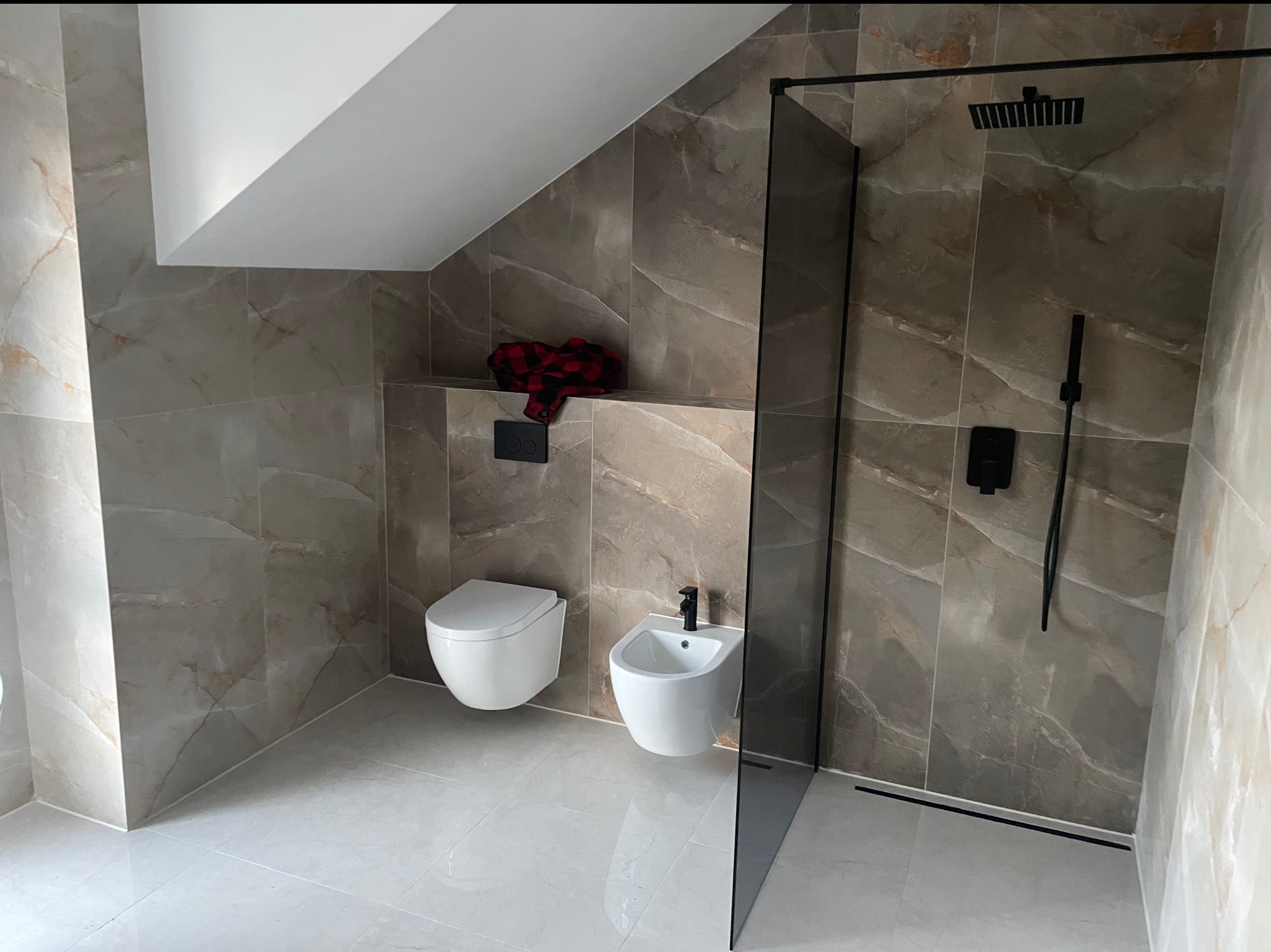 Wykończona łazienka w beżowych płytkach imitujących marmur, z kabiną prysznicową typu walk-in z czarną armaturą i wiszącą toaletą oraz bidetem. Na półce leży czerwona koszula.