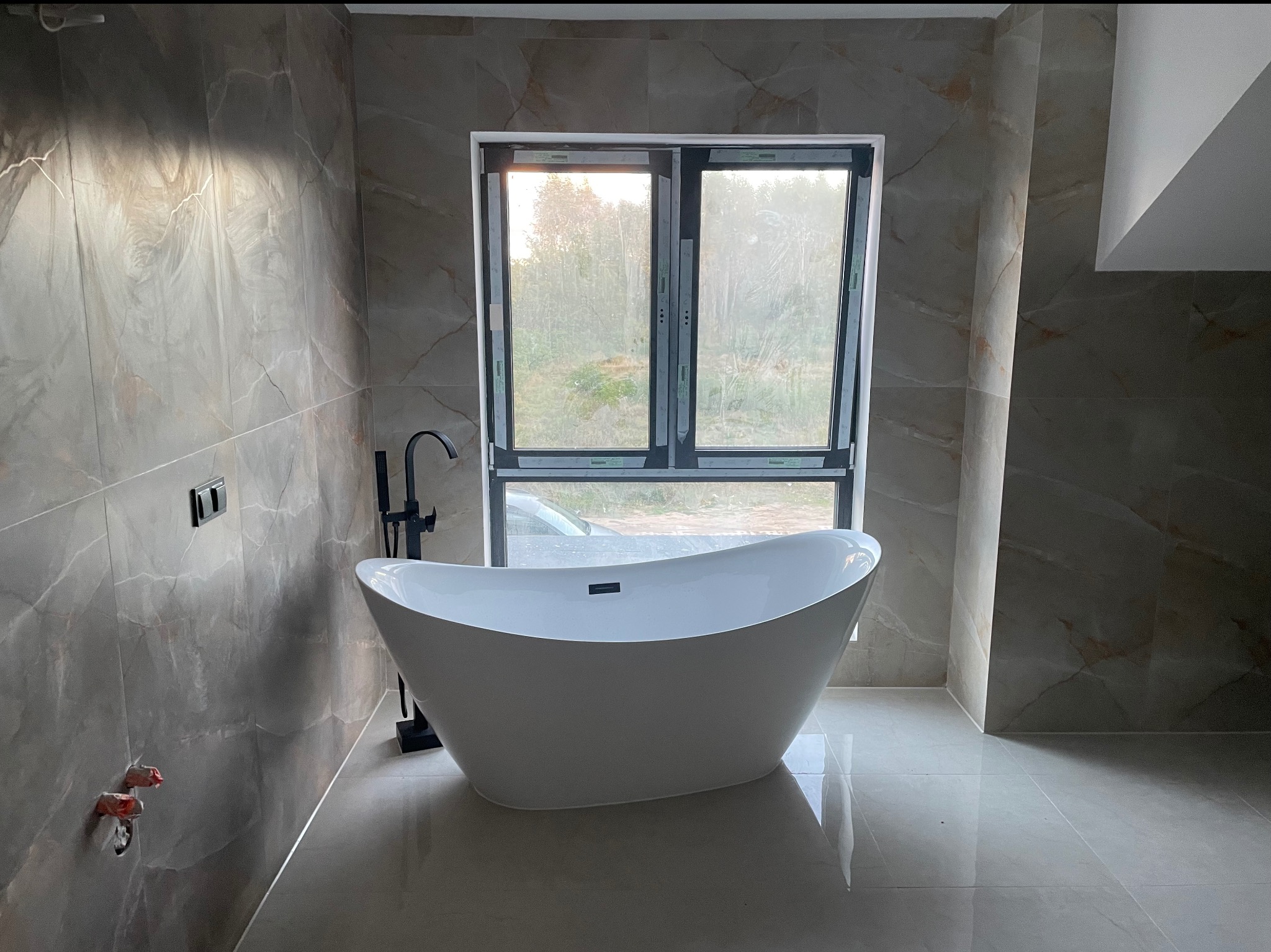 Nowoczesna, wolnostojąca wanna z czarną armaturą na tle okna w wykończonej łazience z szarymi płytkami imitującymi marmur. Widoczne przyłącza wodne.