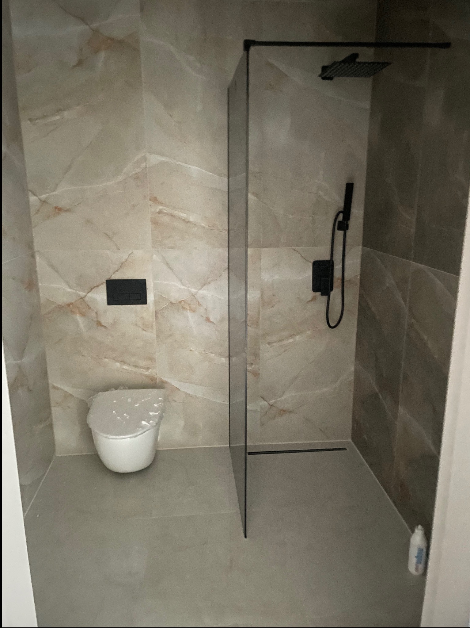 Nowoczesna łazienka z beżowymi płytkami imitującymi marmur, czarną armaturą i kabiną prysznicową typu walk-in. Umywalka z przykryciem ochronnym.