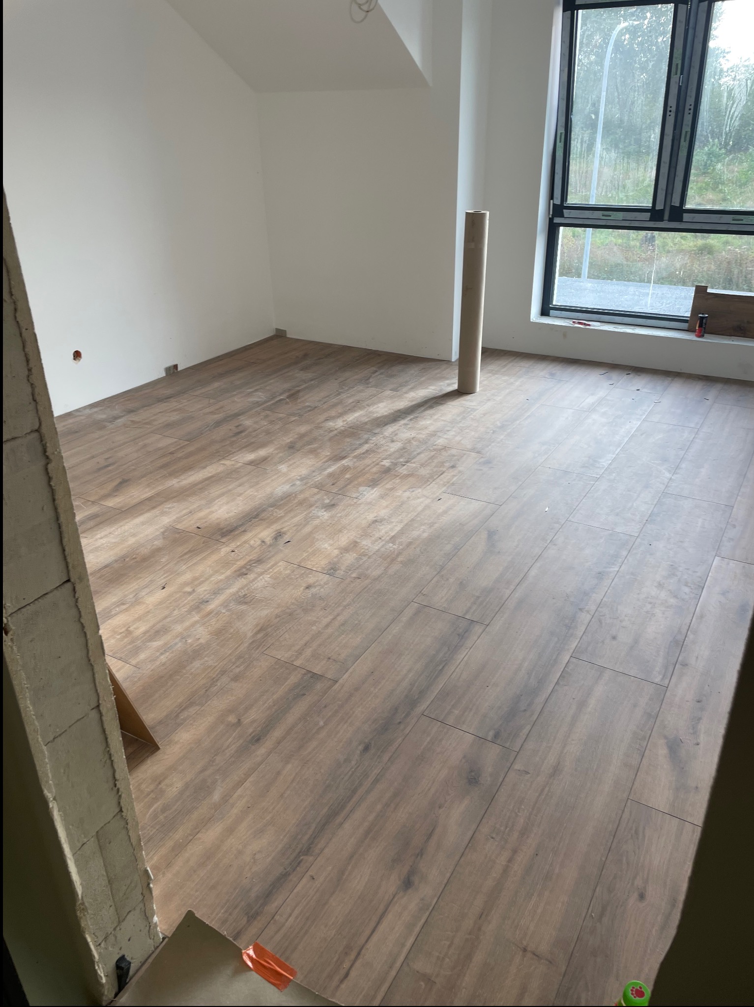 Ukończona podłoga z paneli imitujących drewno w jasnym pokoju z dużym oknem. Widoczny fragment surowej ściany i rolka materiału zabezpieczającego.