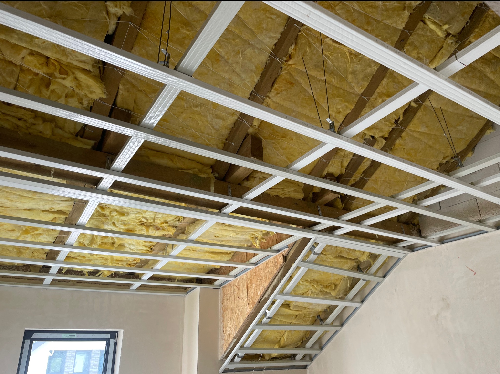 Sufit podwieszany z izolacją z wełny mineralnej, widoczne profile metalowe i drewniana konstrukcja dachu. Ujęcie z dołu, w trakcie montażu.