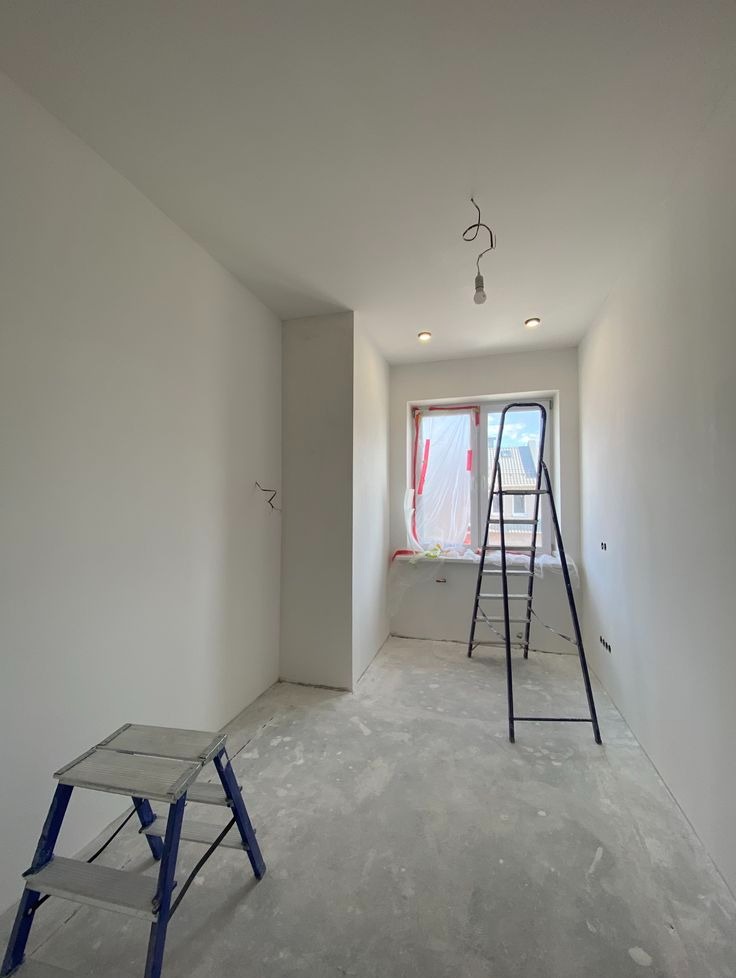 Wnętrze pokoju po świeżym malowaniu na biało, z drabinami i zabezpieczonym oknem. Widoczne ślady po pracach na podłodze i wystające kable. Minimalistyczny styl.