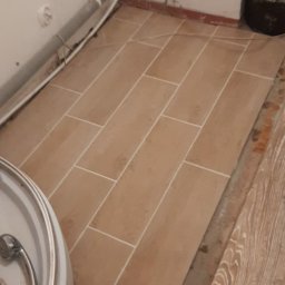 Andrzej Marciniak - Ułożone podłużne, beżowe płytki podłogowe w łazience, widoczne rury przy ścianie i fragment kabiny prysznicowej. Widoczne niedociągnięcia przy łączeniu z innym rodzajem podłogi.