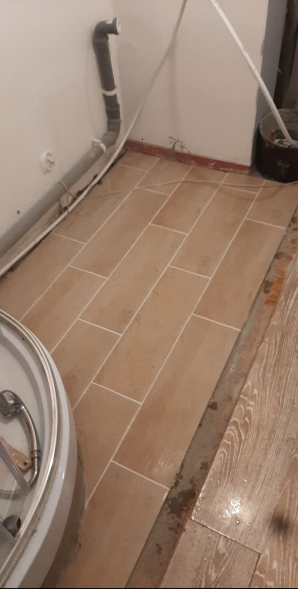 Ułożone podłużne, beżowe płytki podłogowe w łazience, widoczne rury przy ścianie i fragment kabiny prysznicowej. Widoczne niedociągnięcia przy łączeniu z innym rodzajem podłogi.