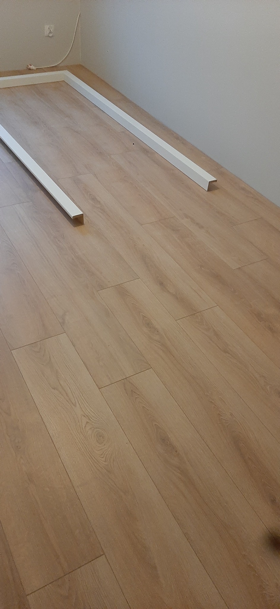 Jasne panele podłogowe z zamontowanymi białymi listwami przypodłogowymi wzdłuż ścian. Widok z góry, ukośny kadr.