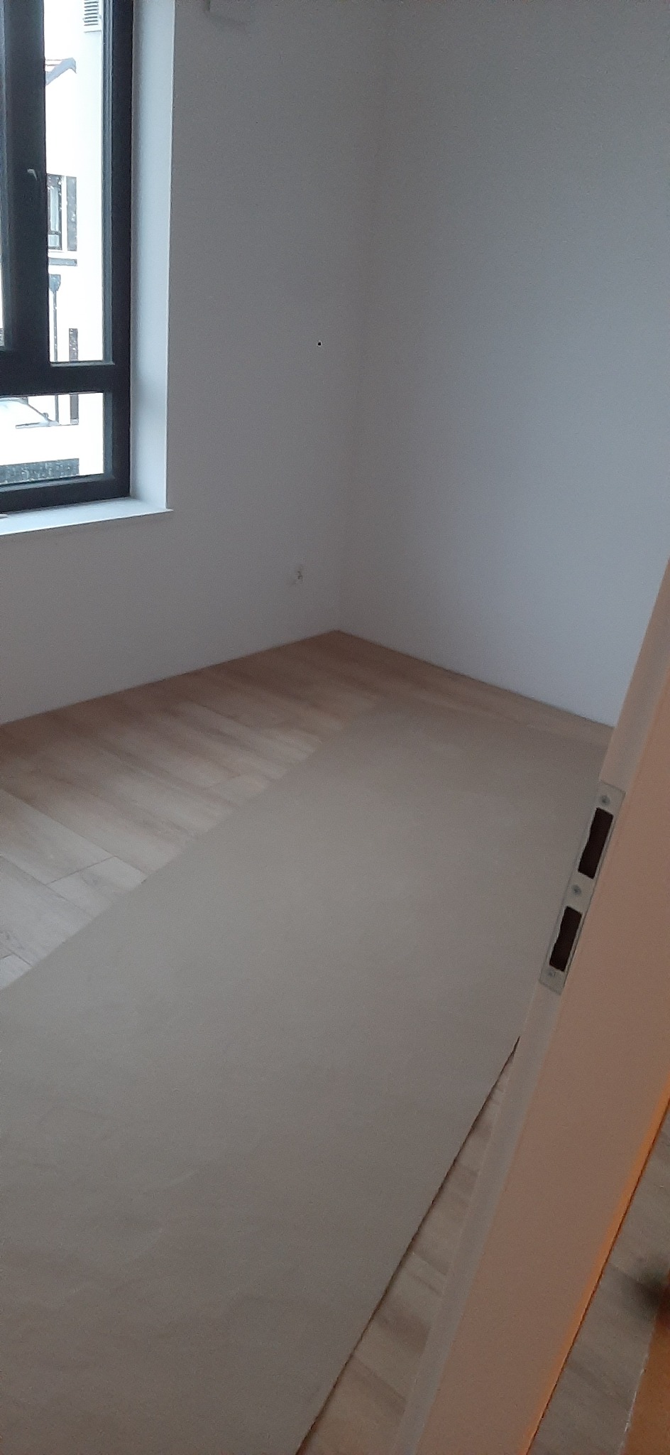 Pokój po remoncie z jasną podłogą zabezpieczoną kartonem falistym. Widoczne okno i fragment białych drzwi z zamkiem. Minimalistyczne wnętrze.