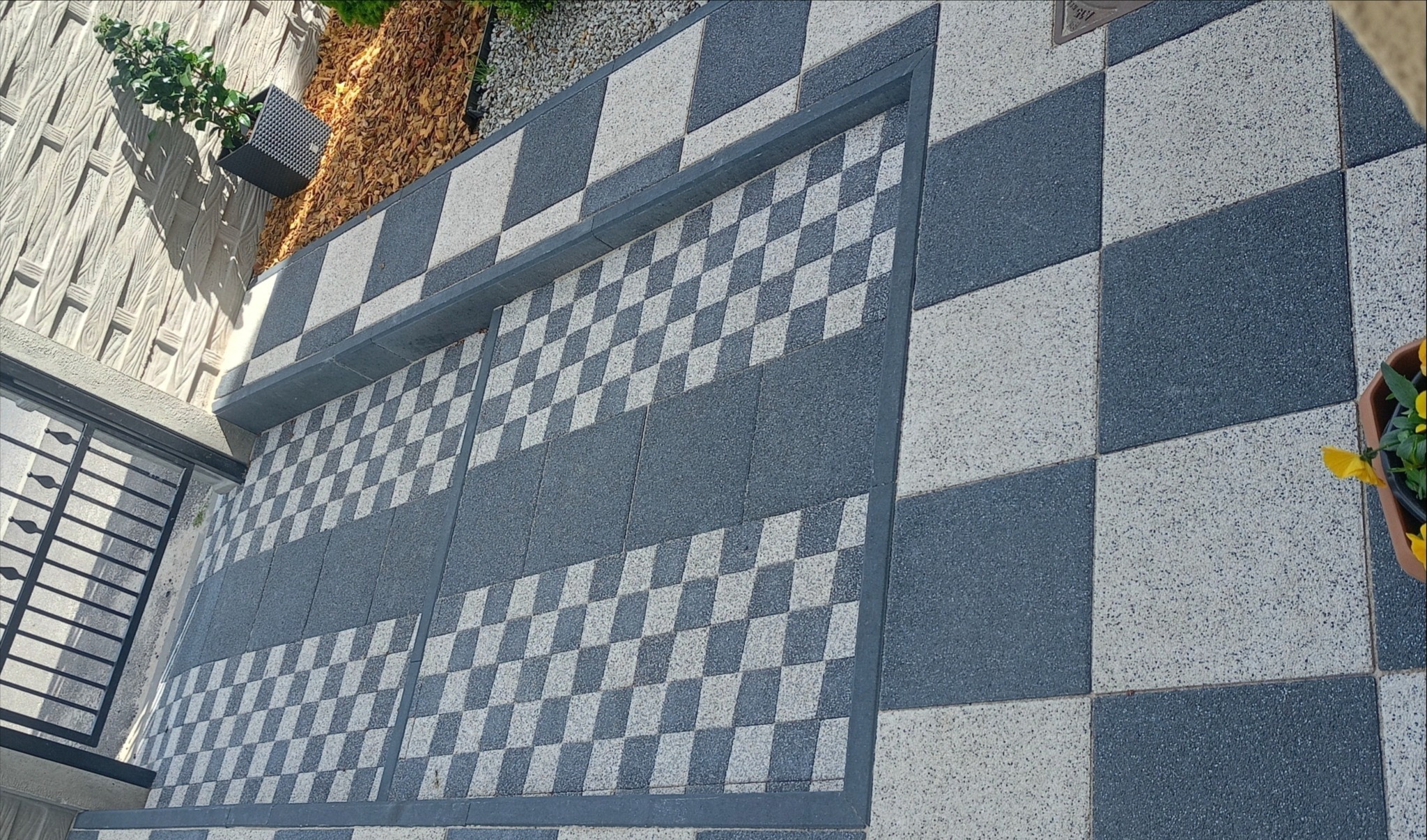 Szachownica z kostki brukowej w odcieniach szarości, z doniczką z kwiatami w rogu. Ujęcie z góry, widoczny fragment ogrodzenia i roślinność.